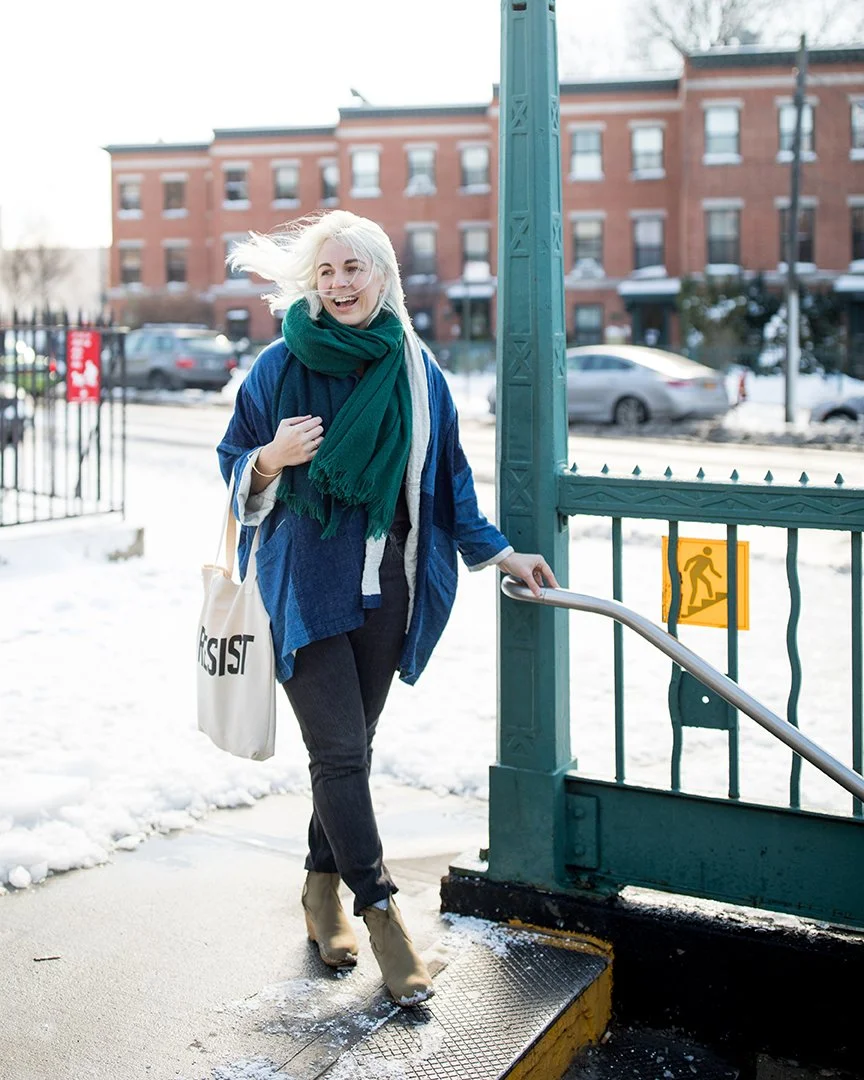 A woman with blonde hair, wearing a green scarf, blue jacket, and black pants, is smiling and holding onto a handrail as she exits a subway station in winter. Snow is on the ground and in the background, with some houses and cars visible.