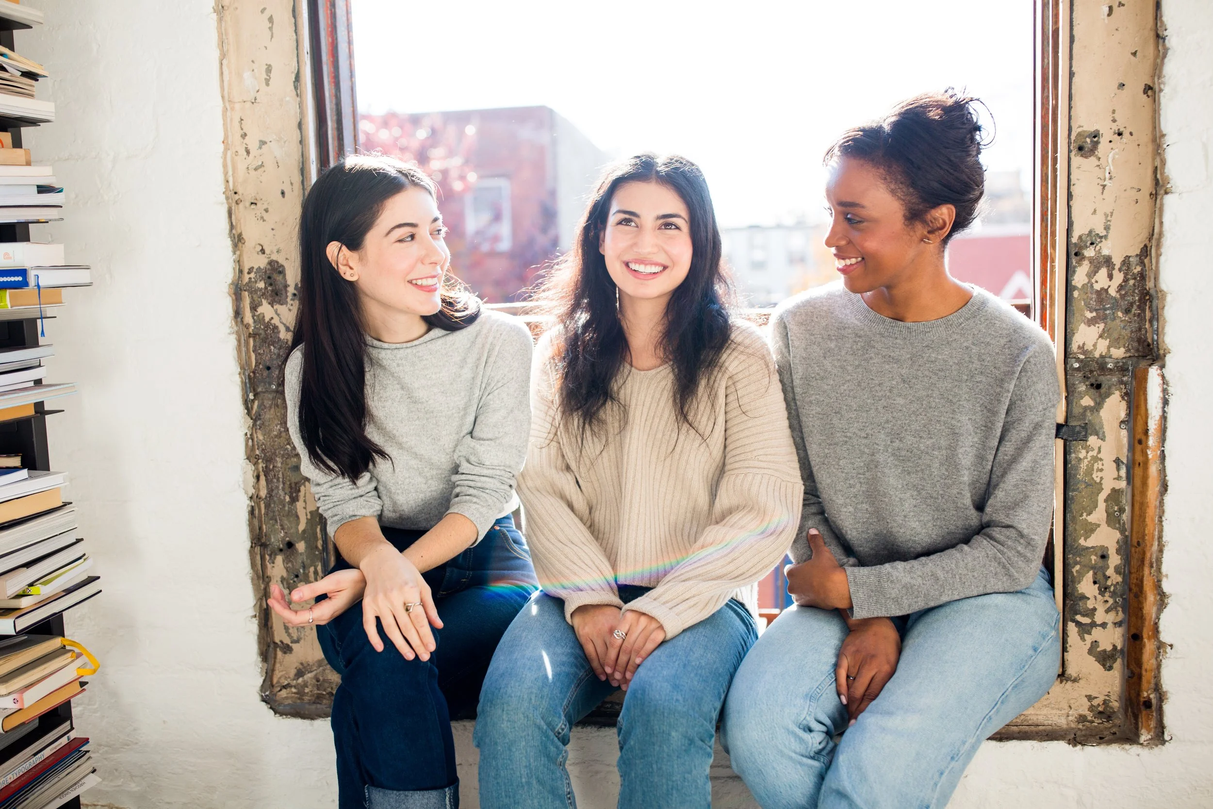 Three women sitting on a windowsill engaged in conversation, with a bookshelf on the left and a cityscape visible through the window.