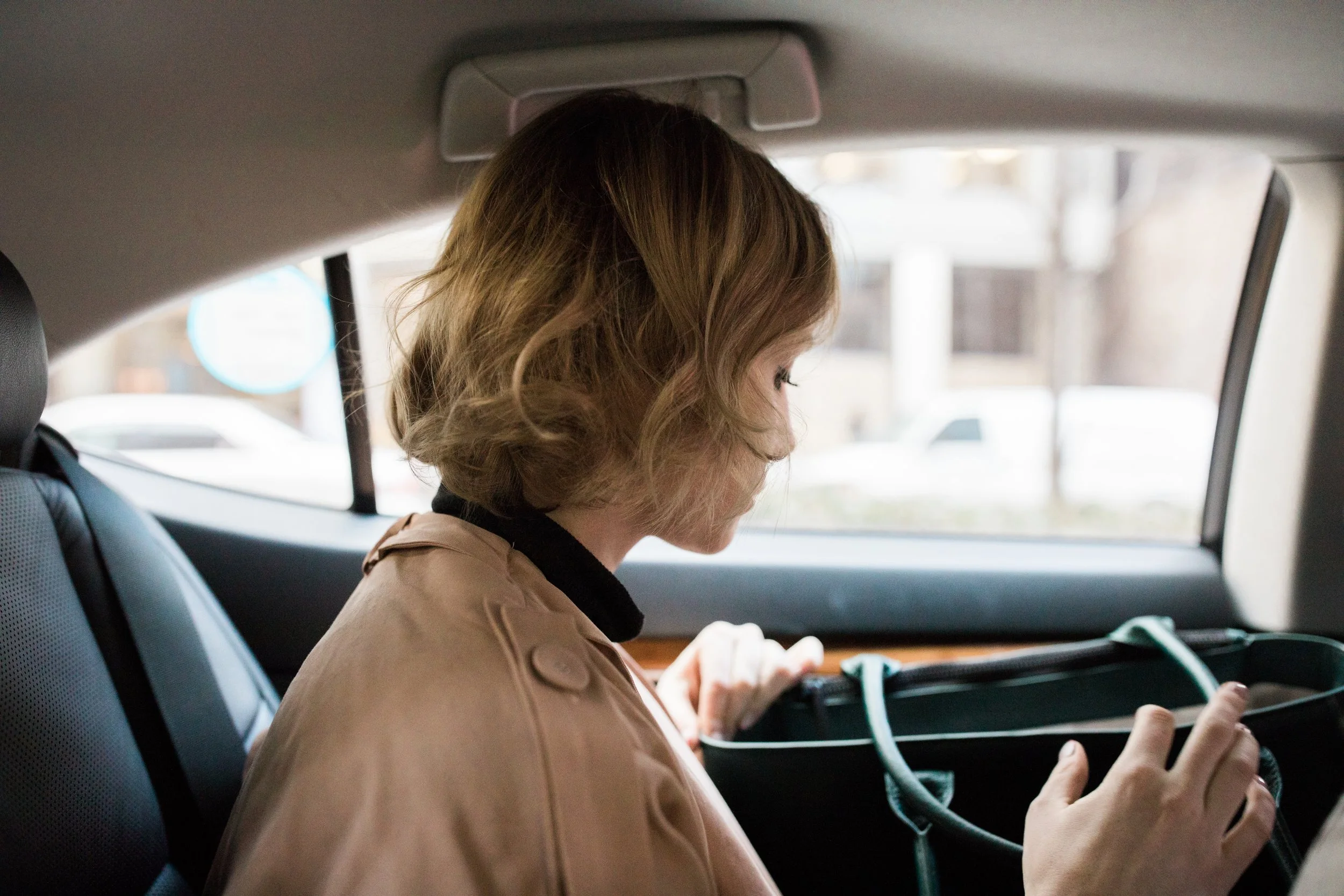 A woman with curly, shoulder-length hair sitting in the backseat of a car, looking at her phone. She is wearing a beige jacket and is near the window, with a blurred cityscape outside.