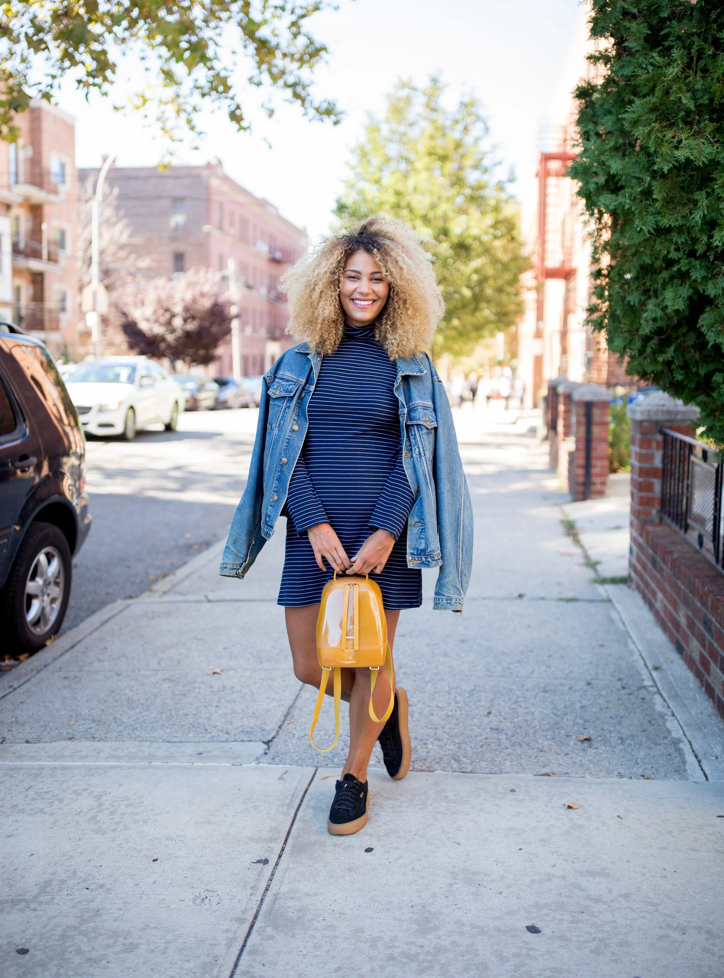 A young woman with curly blonde hair, wearing a denim jacket and a navy striped dress, walking on a city sidewalk with a yellow backpack. She is smiling and looking at the camera, with buildings and trees in the background on a sunny day.