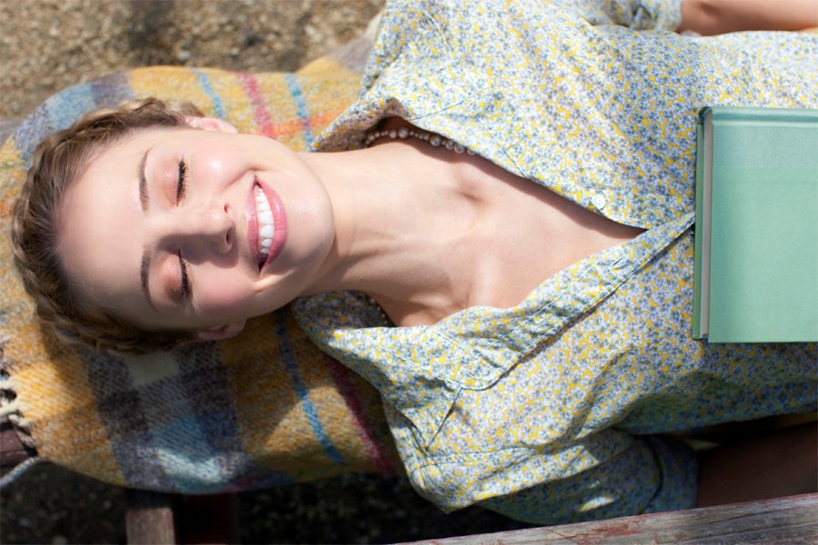 A woman with closed eyes and a big smile lying on a colorful blanket with a light blue book nearby.