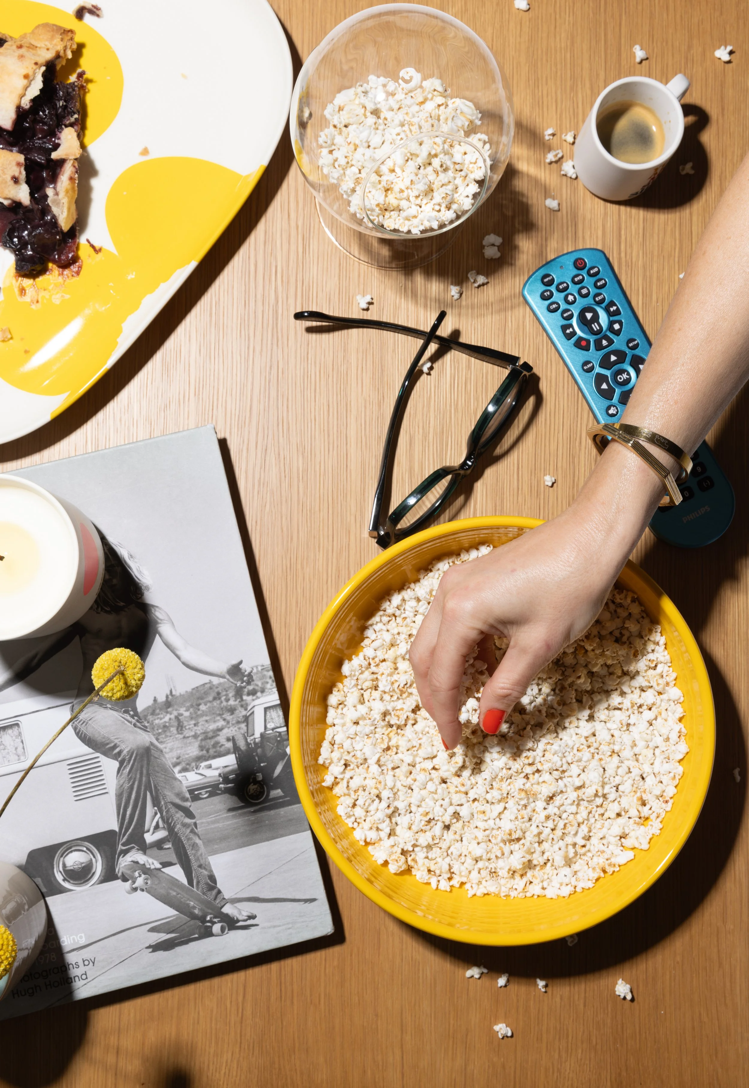 Person's arm and hand reaching into a yellow bowl of popped popcorn on a wooden table, with a black-framed pair of glasses, a remote control, a coffee mug, a glass bowl of popcorn, a black and white magazine photo, a cup of coffee, and a slice of blueberry pie on a white plate visible on the table.