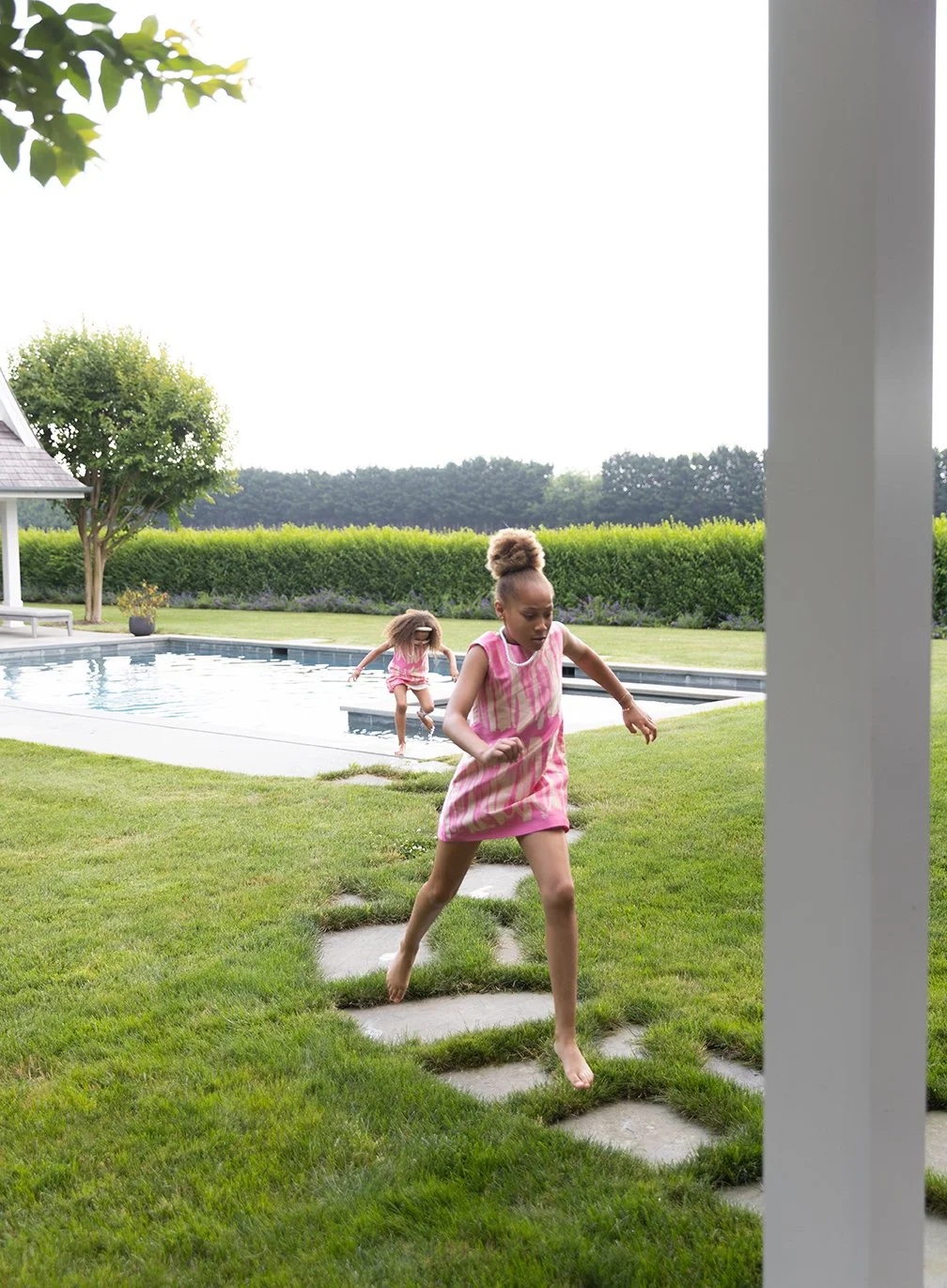 Two young girls playing by the swimming pool, one running on stepping stones on the grass in the foreground, the other near the pool in the background.