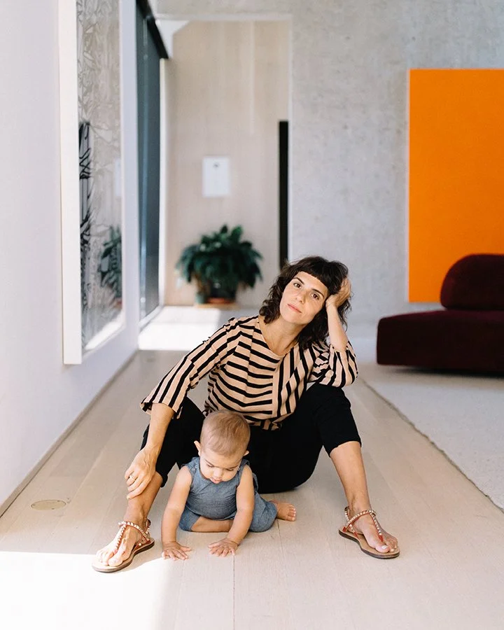 A woman and a young child sitting on a wooden floor in a modern, brightly lit room. The woman, wearing a striped top and black pants, sits with her legs apart, one hand on her head and the other on the child's back. The child, dressed in a denim outfit, is crawling on his hands and knees between her legs. The room has contemporary decor with a large orange and gray wall, a red couch, and a potted plant in the background.