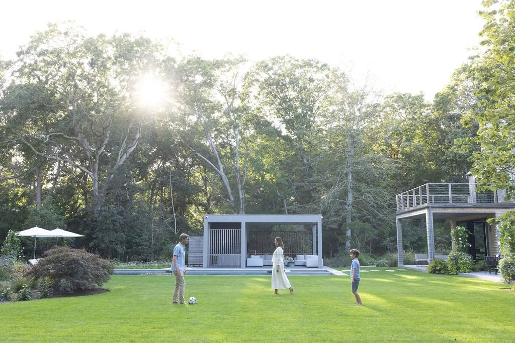 Family playing soccer on a lush green backyard with modern house and outdoor seating area, surrounded by trees.