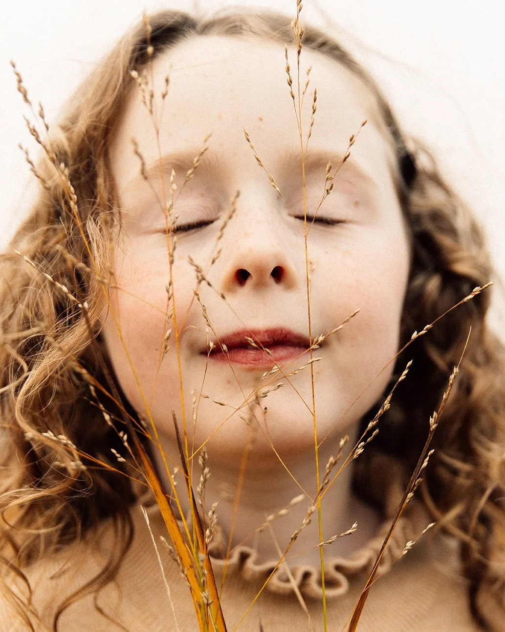 Close-up of a young girl with curly hair softly smiling with her eyes closed, holding dried grass in front of her face.