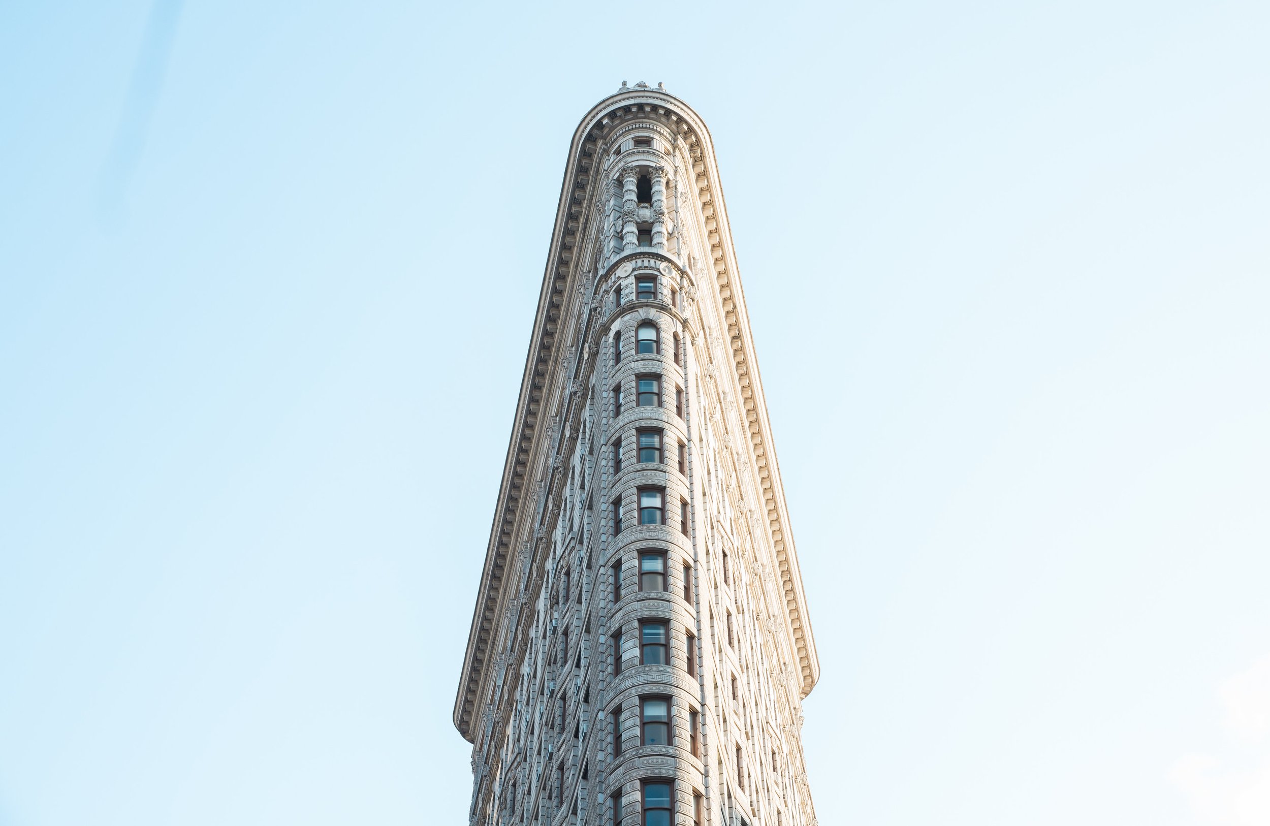 A tall, ornate skyscraper, known as the Flatiron Building, with intricate stone detailing and multiple windows, against a clear blue sky.