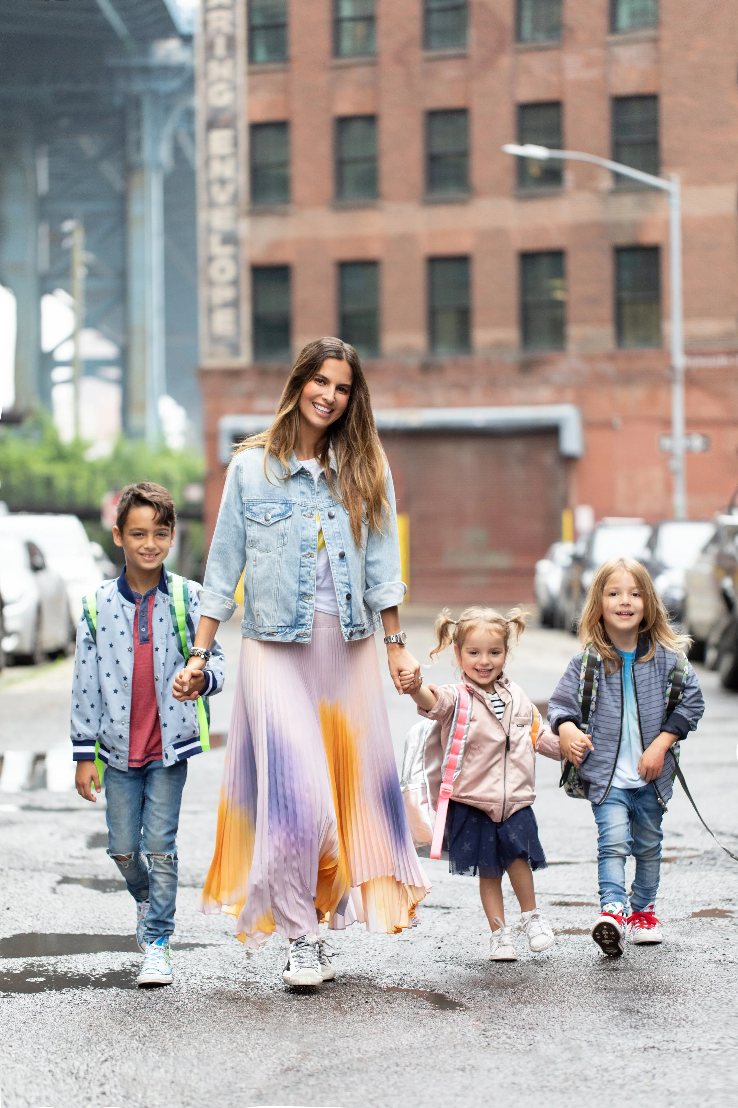A woman holding hands and walking with five children on a city street with buildings and parked cars in the background.