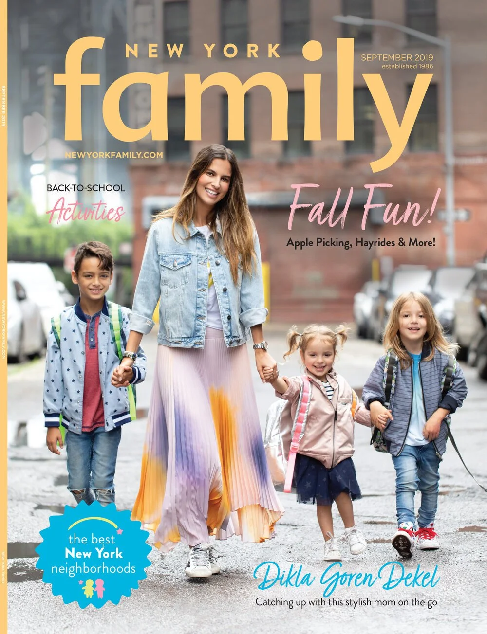A woman and three children walking on a street in New York City, holding hands. The woman is smiling and wearing a denim jacket and a colorful pleated skirt. The children are school-aged, wearing backpacks, and smiling. Cars are parked along the stre