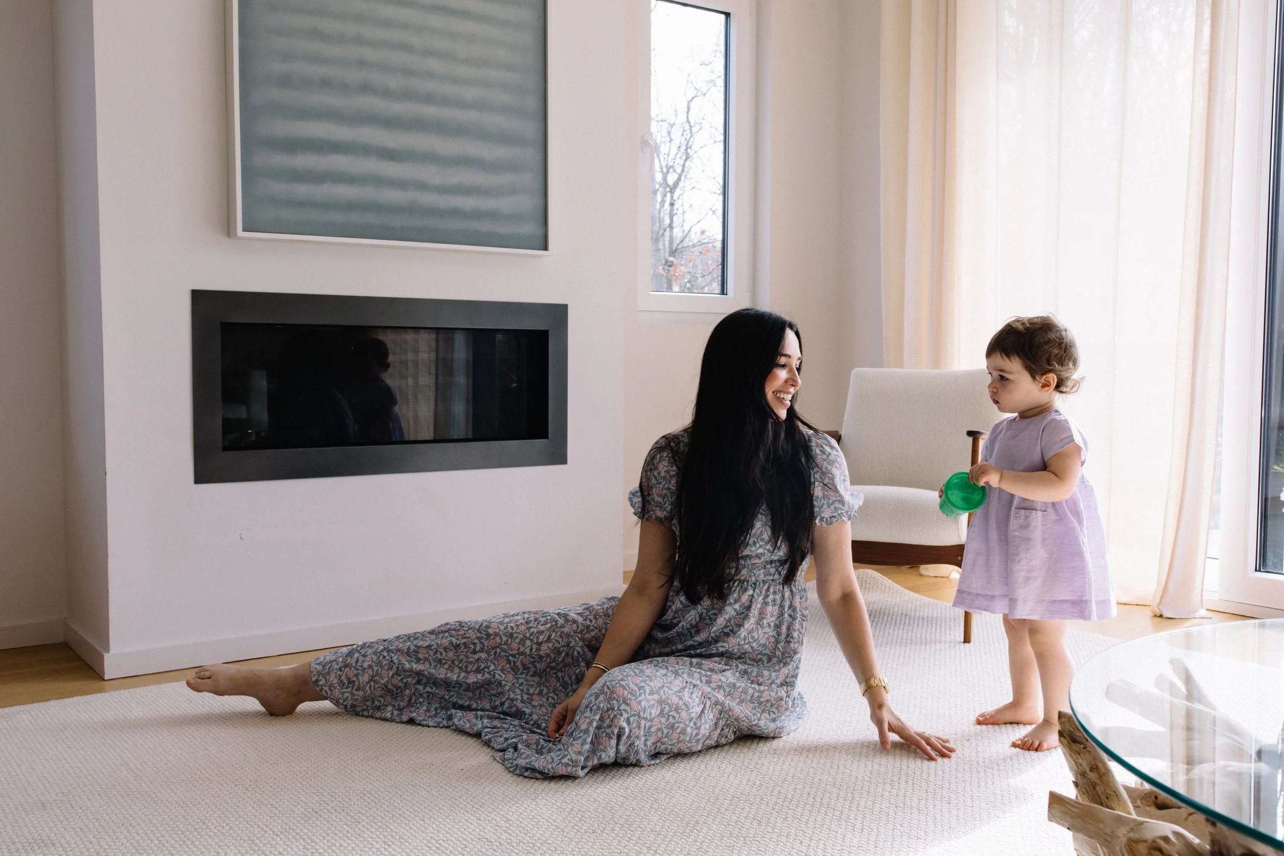 A woman with long dark hair, wearing a floral dress, sitting on a beige carpet with her legs stretched out, smiling at a young girl, who is standing and holding a green toy, in a bright living room with a fireplace, a large window, and cream-colored 