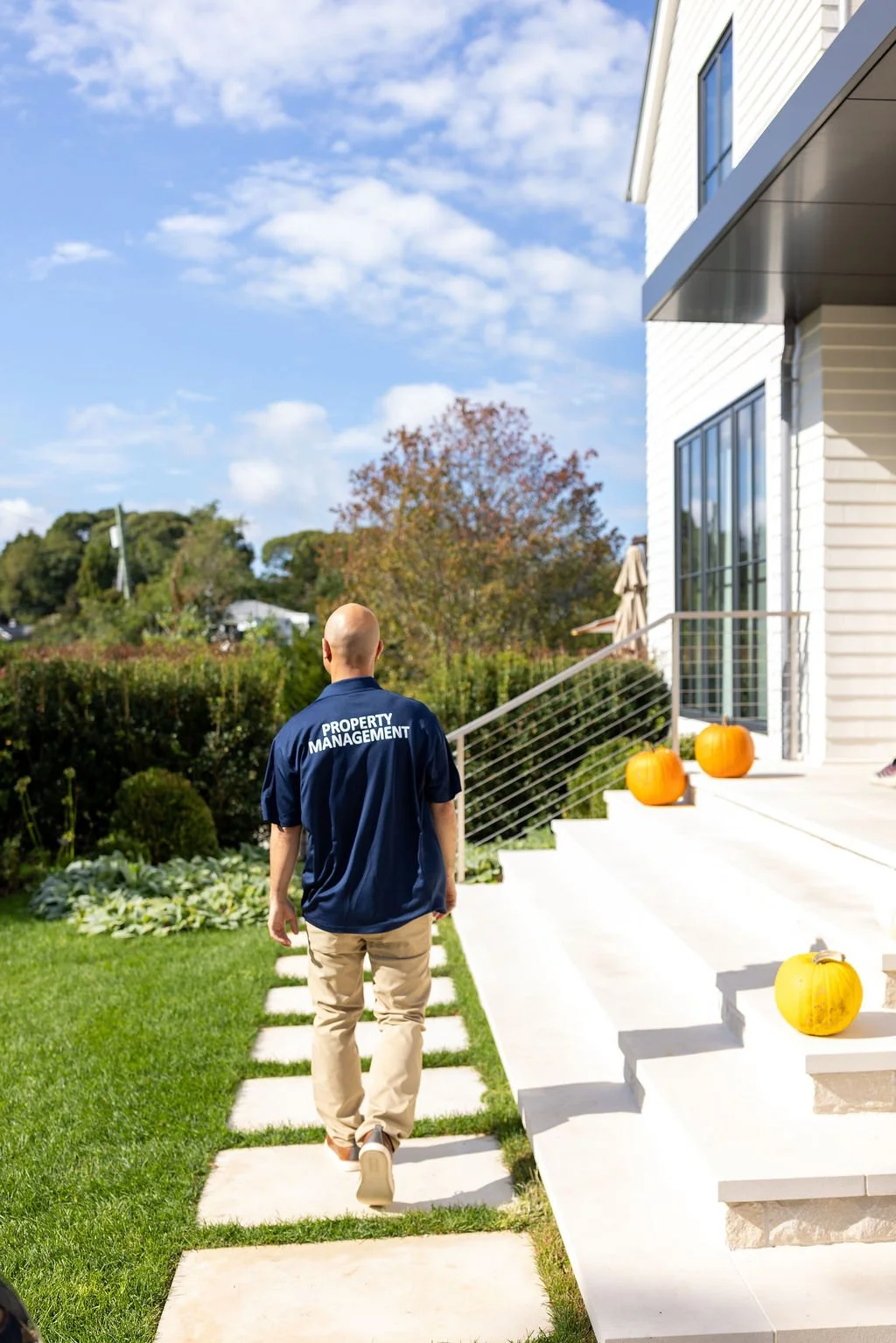 A property manager walking towards a modern white house with pumpkins on the steps, a manicured lawn, and trees in the background on a sunny day with a partly cloudy sky.