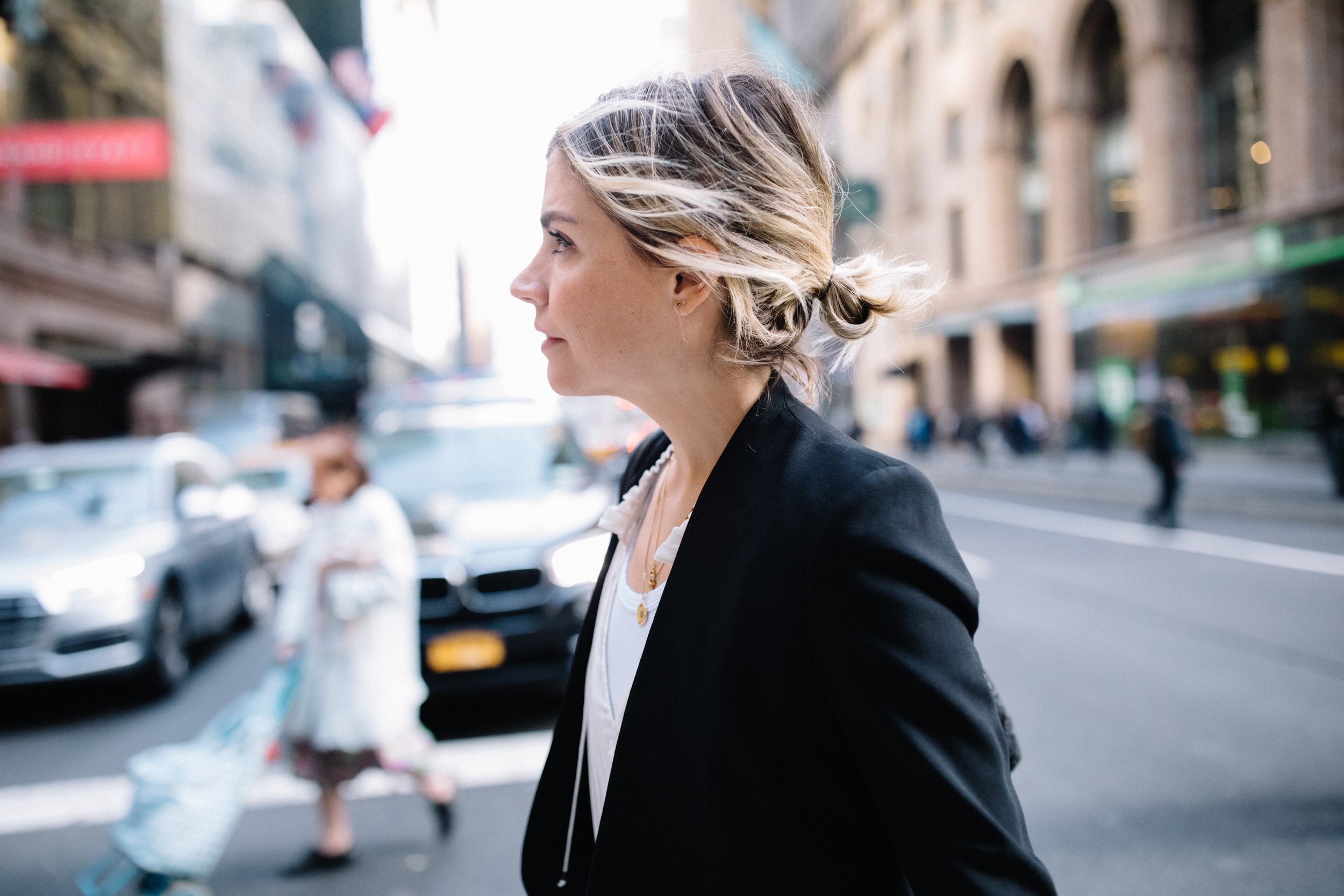 Side profile of a woman with blonde hair tied back, walking on a city street.