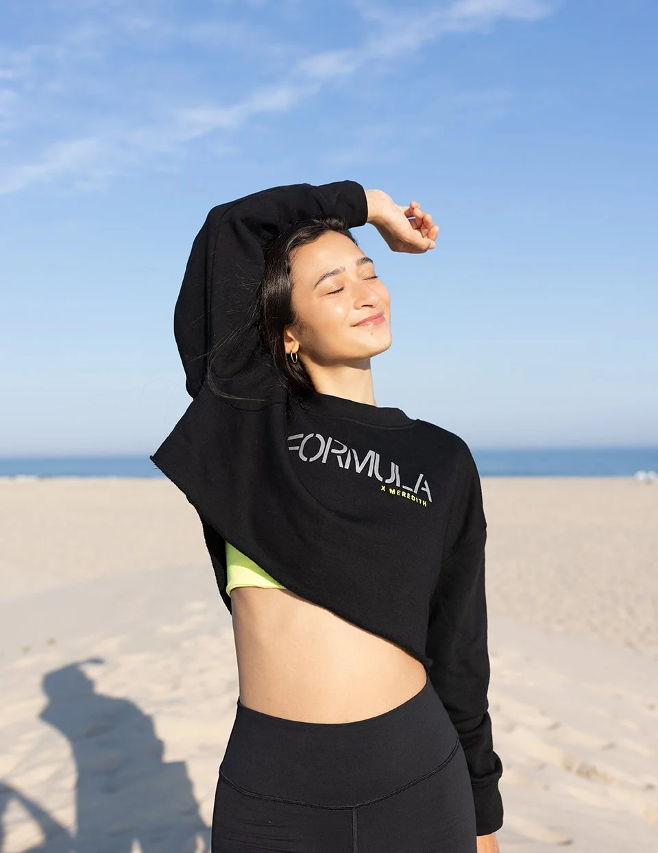 Young woman with dark hair smiling with eyes closed, wearing a black sweatshirt with the word 'FORMULA' and high-waisted black leggings, standing on a beach with sand, blue sky, and ocean in the background.