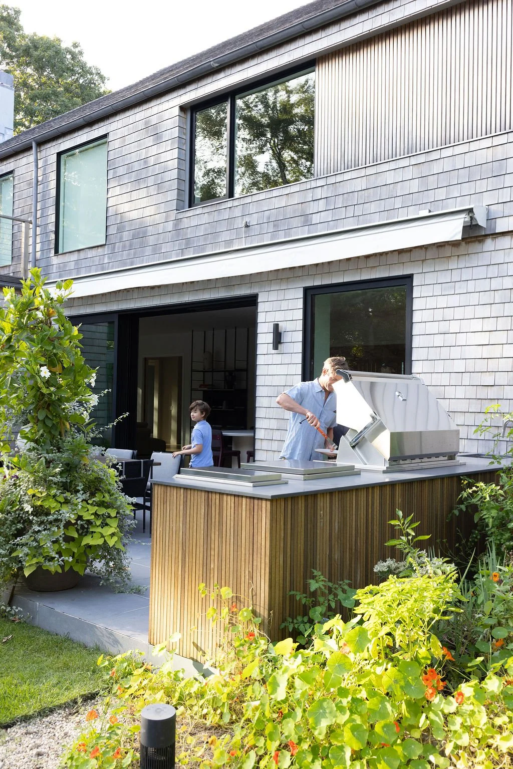 A man grilling food on a stainless steel outdoor barbecue at a modern house with large windows, surrounded by lush green plants and garden flowers, with a young boy standing nearby.