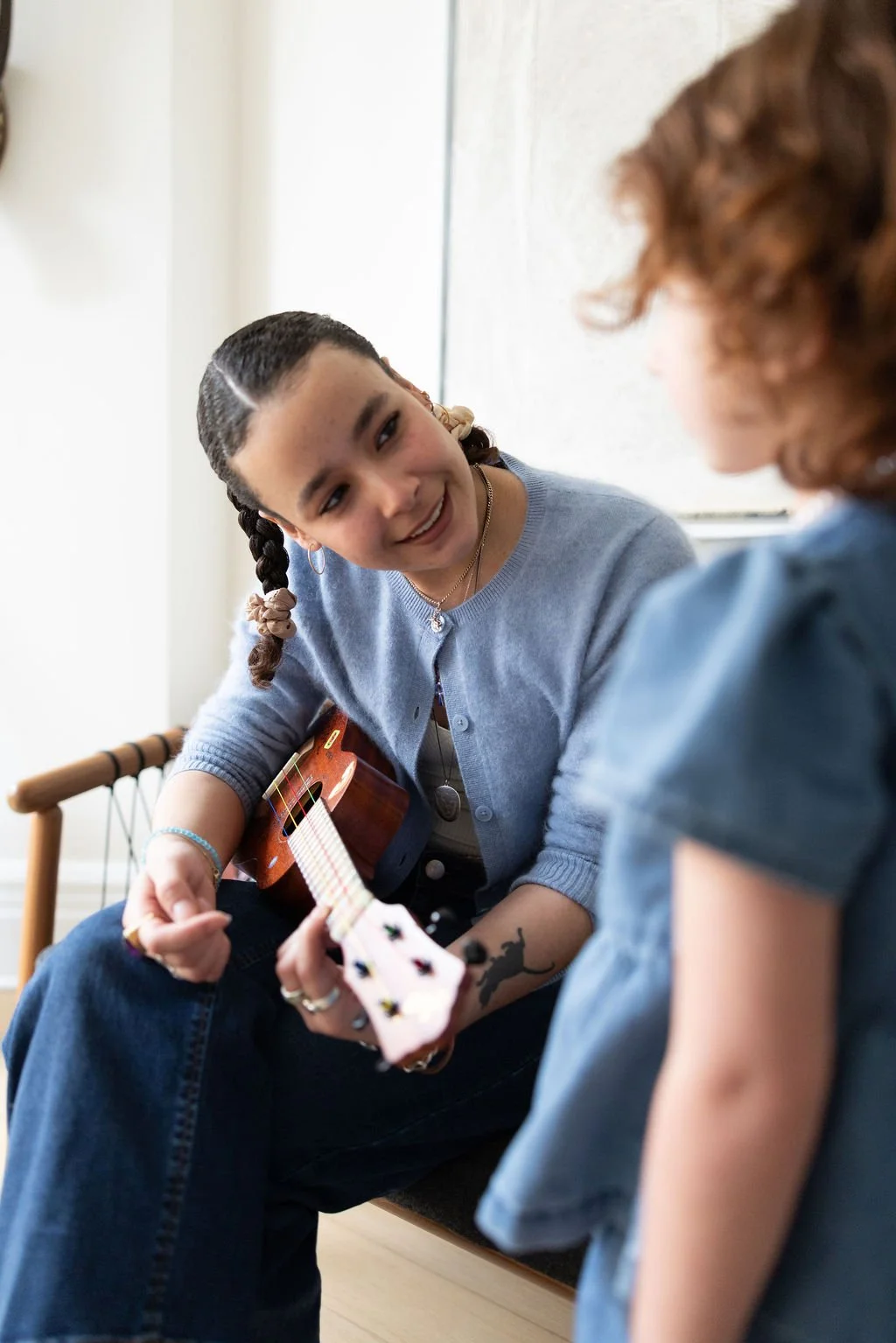 A young woman with braided hair playing a guitar and smiling at a child with curly hair.