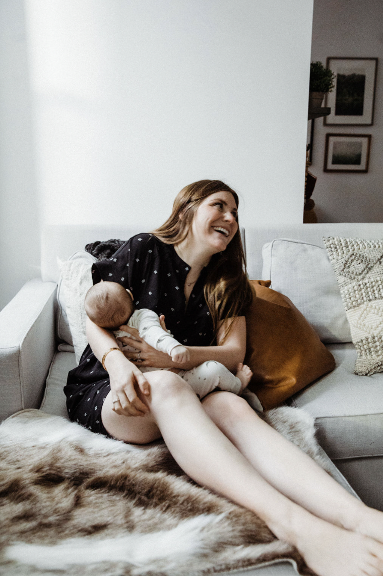 A woman with long brown hair, wearing a black polka dot dress, sitting on a sofa with a baby in her arms, smiling and looking up. The sofa has pillows and a faux fur blanket, in a cozy living room with framed pictures on the wall.