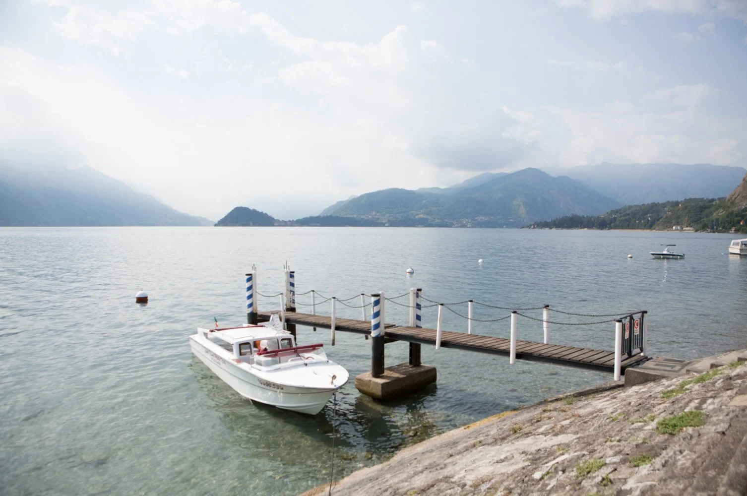 A tranquil lake with boats, mountains in the background, a wooden dock extending into the water, and a cloudy sky.