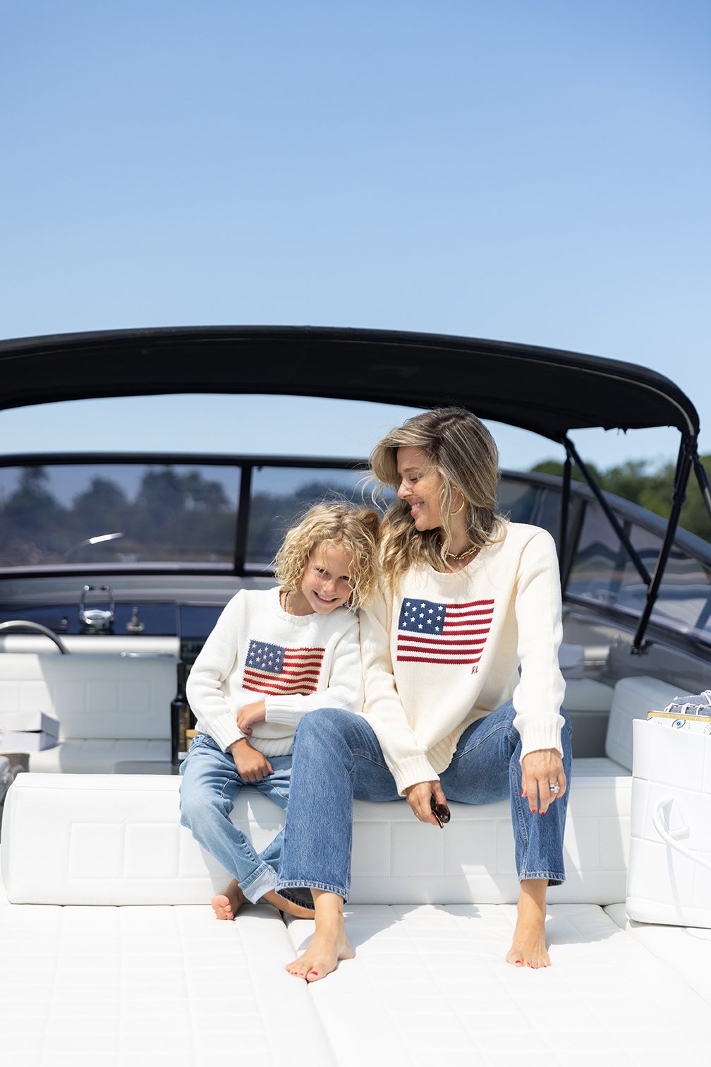 A woman and girl sitting on a white boat, both wearing matching cream sweaters with an American flag on the front, smiling and looking at each other.