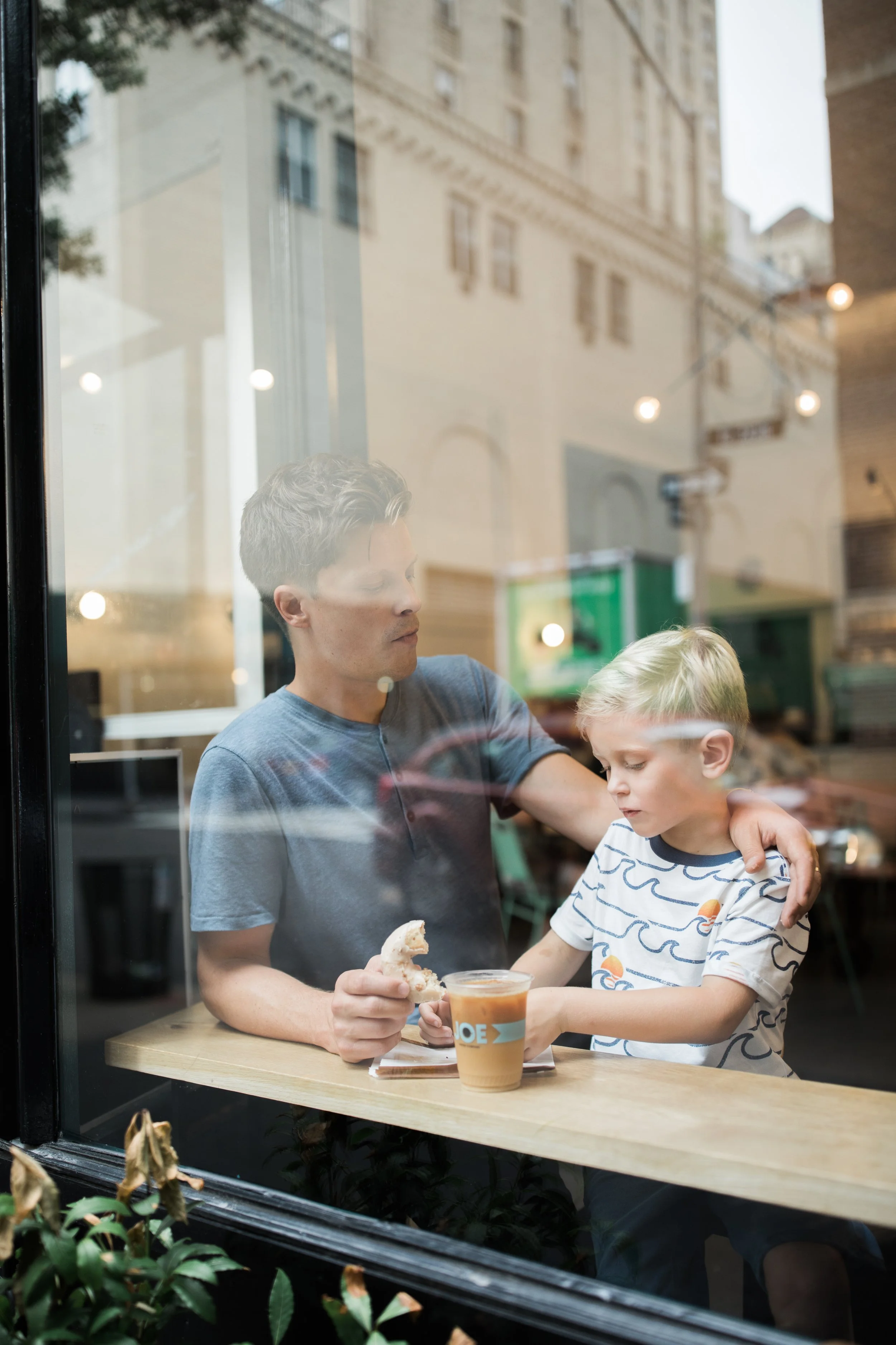 A young man with curly blond hair and a young boy with blond hair and glasses sit at a wooden table inside a cafe, seen through a window. The man holds a sandwich, and the boy has a cup of orange juice.