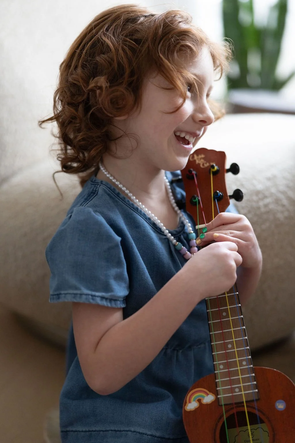 A young girl with curly red hair smiling happily while holding a small colorful ukulele.