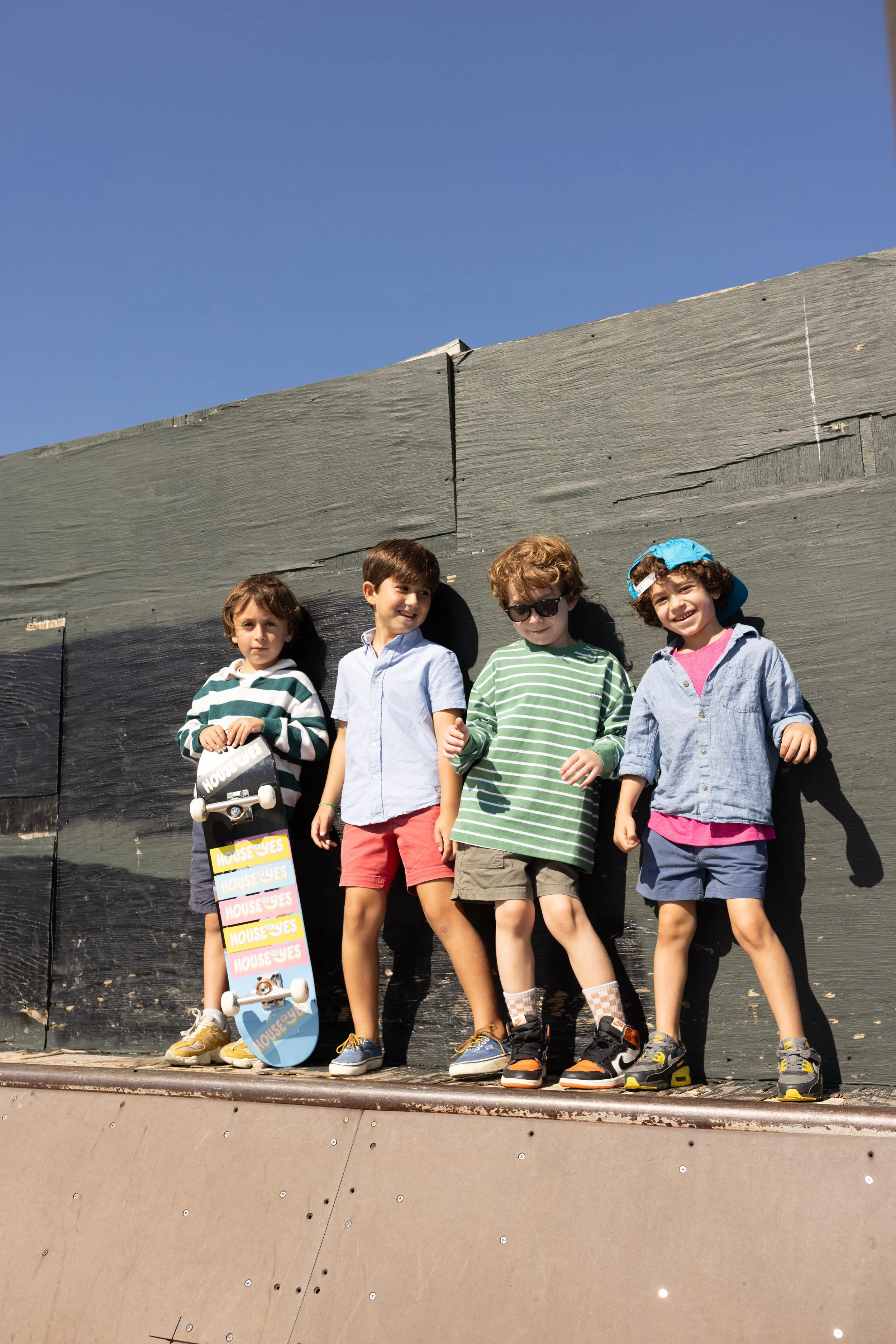 Four kids standing on a skateboard ramp against a gray wall with a clear blue sky, smiling and posing for the camera.