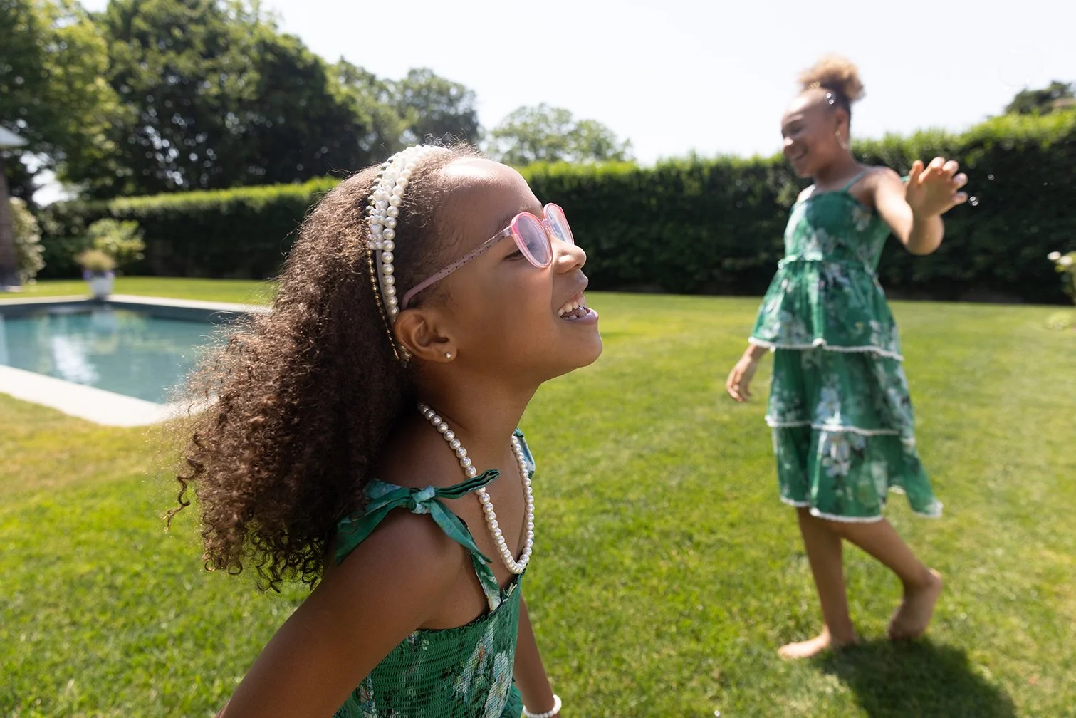 Two young girls in green dresses playing outdoors near a swimming pool, with one girl laughing and the other reaching out her hand, on a sunny day with green trees and grass in the background.