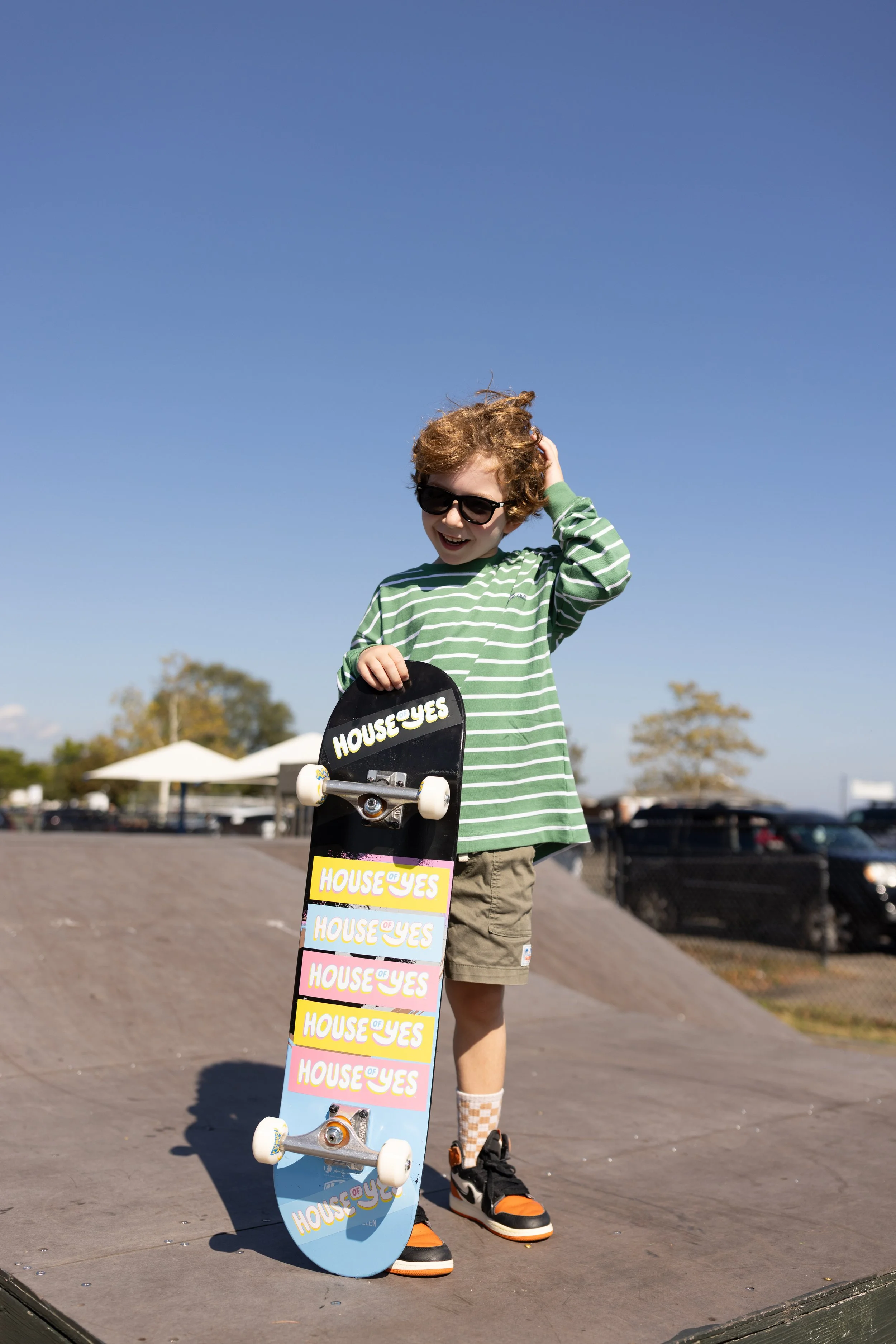 A young boy with curly hair wearing sunglasses, a green striped shirt, beige shorts, and skateboard shoes while holding and standing next to a skateboard with the words 'House of Yes' on it, outdoors on a sunny day with trees and parking lot in the b