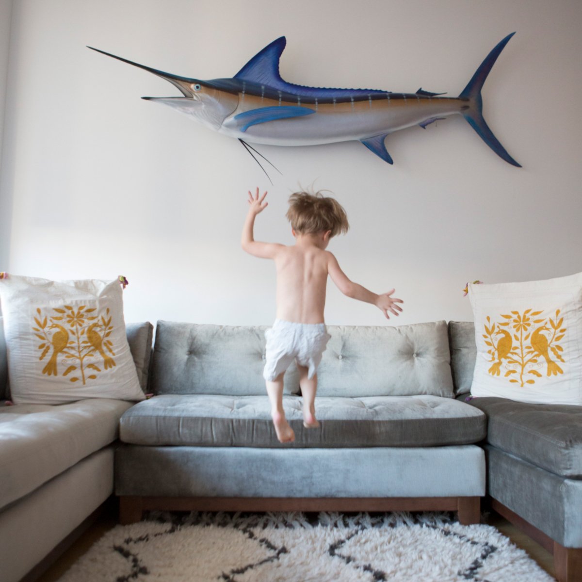 A child jumping on a sofa with a large marlin fish model hanging on the wall above.