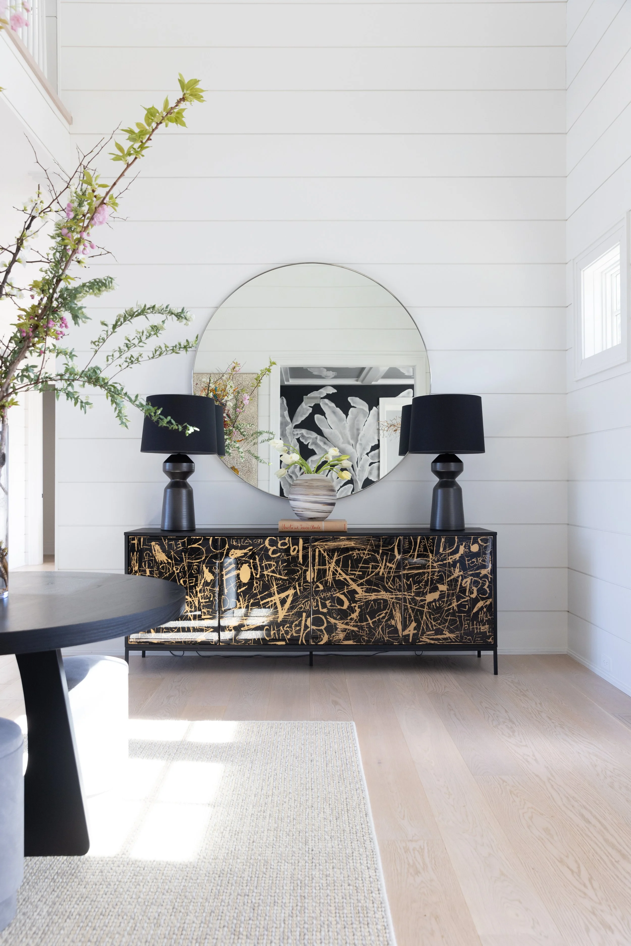 Modern interior with a black console table featuring gold scribble designs, a large round mirror, two black table lamps, a striped vase with white flowers, and a print of white leaves on the wall reflected in the mirror. The room has white shiplap walls and light wood flooring.