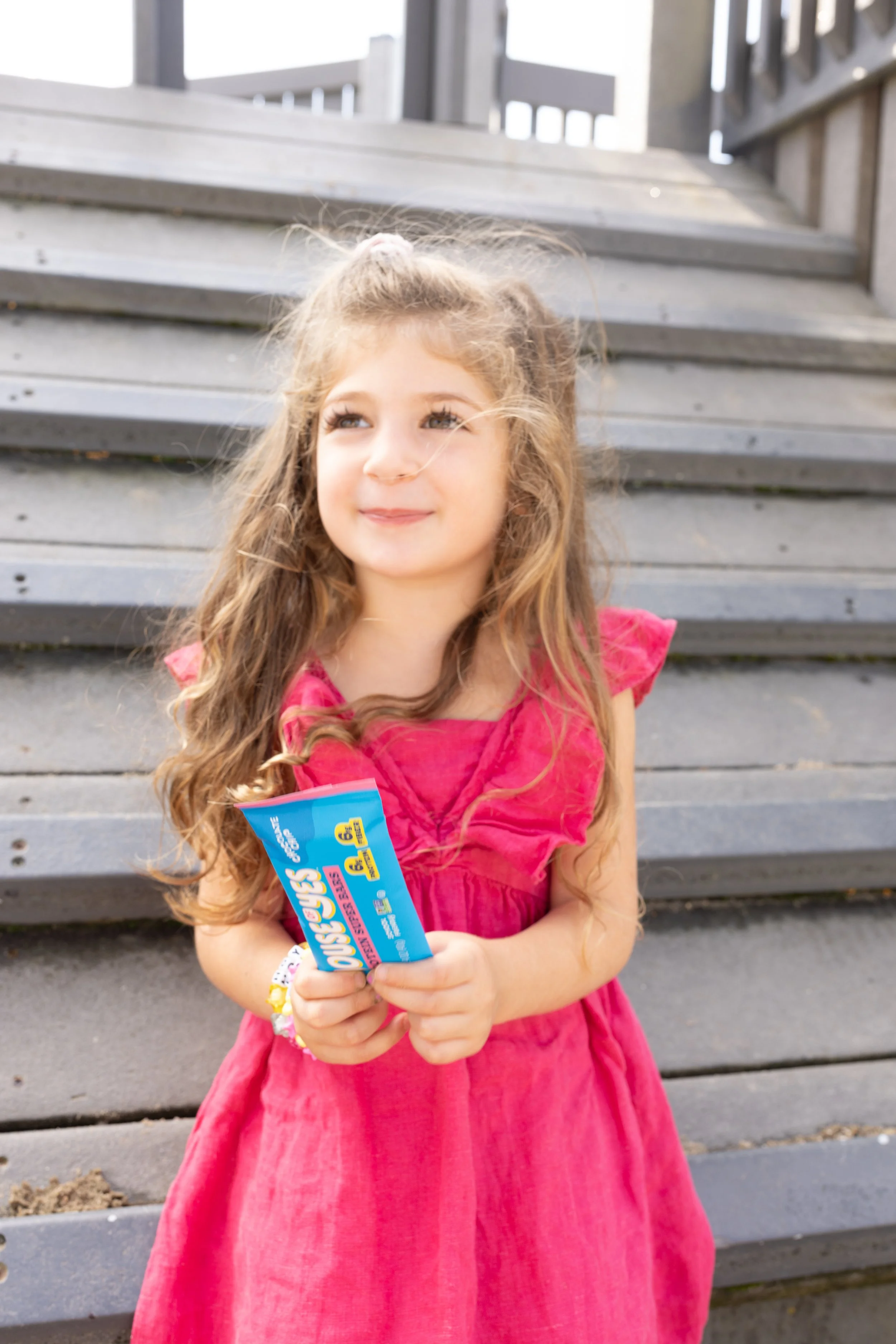 Young girl with long light brown hair wearing a pink dress, holding a blue candy wrapper, standing in front of wooden stairs outdoors.