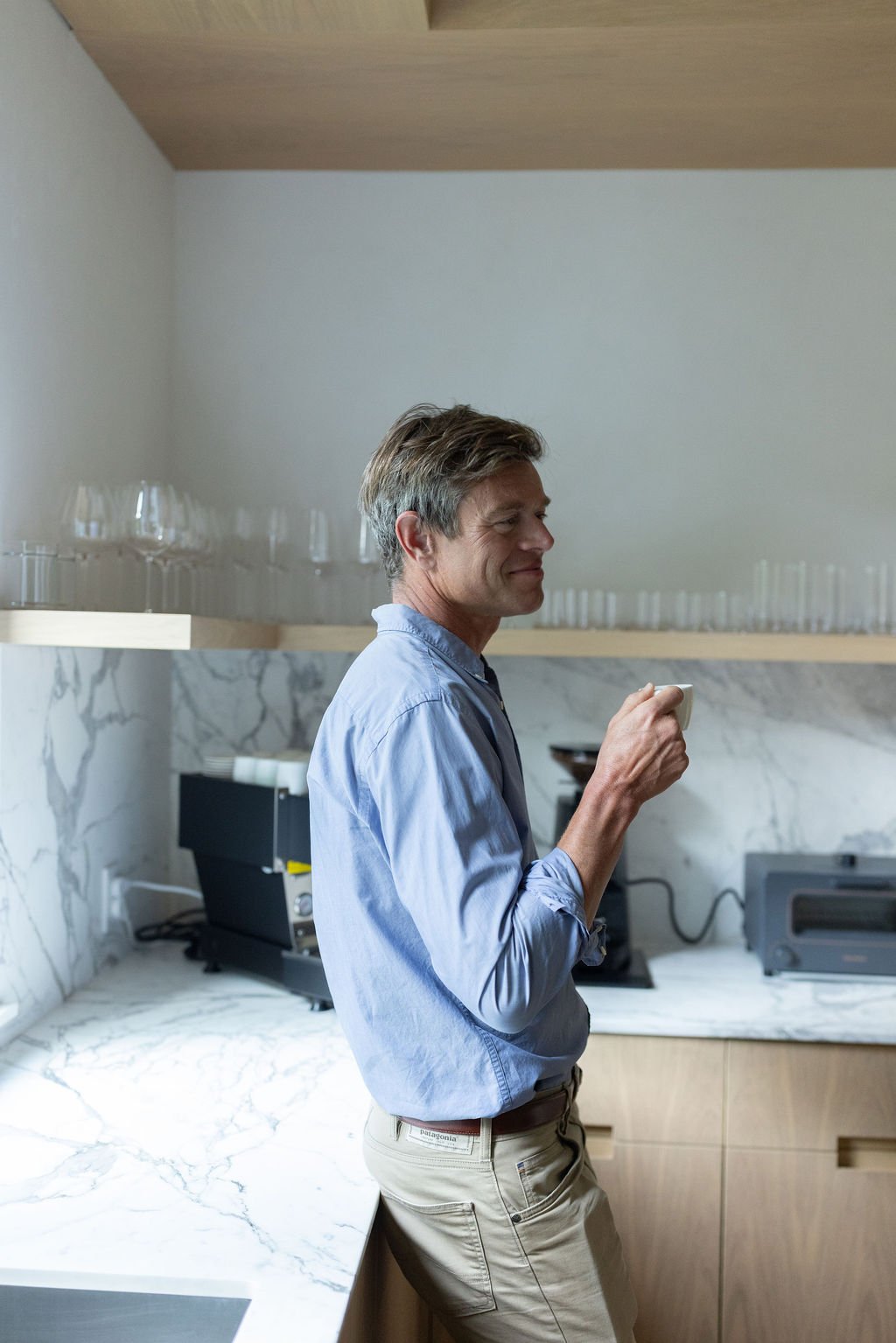 A man standing in a modern kitchen, holding a cup, with glassware on a shelf and kitchen appliances on the counter.