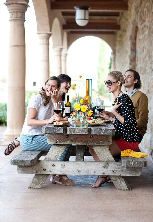 Four people sit around a rustic picnic table outdoors, enjoying wine and conversation. The table has a vase with yellow flowers, wine bottles, and snacks, with a stone building and arches in the background.