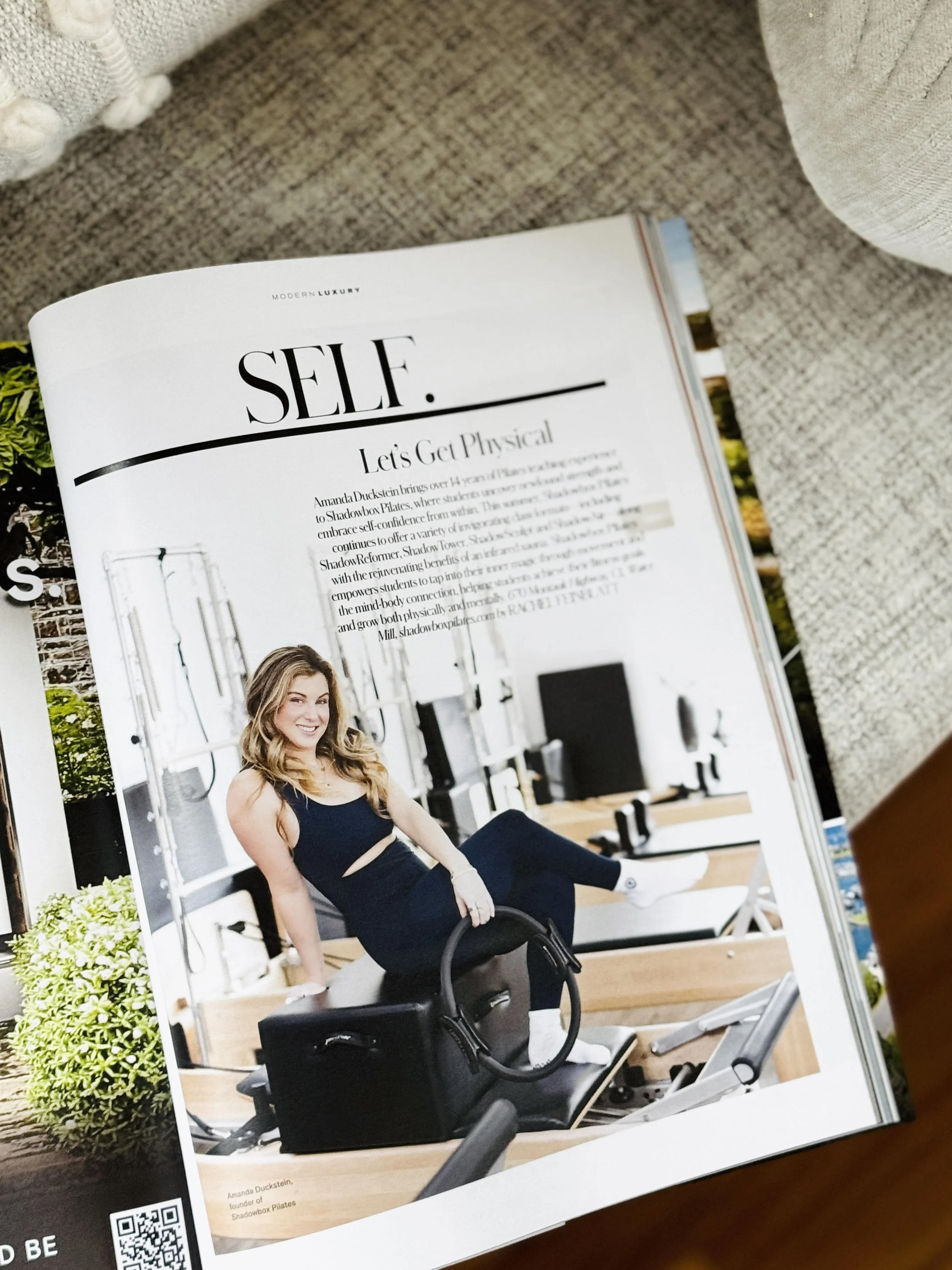 A woman doing Pilates on a reformer machine in a fitness studio, smiling, with greenery outside the window.