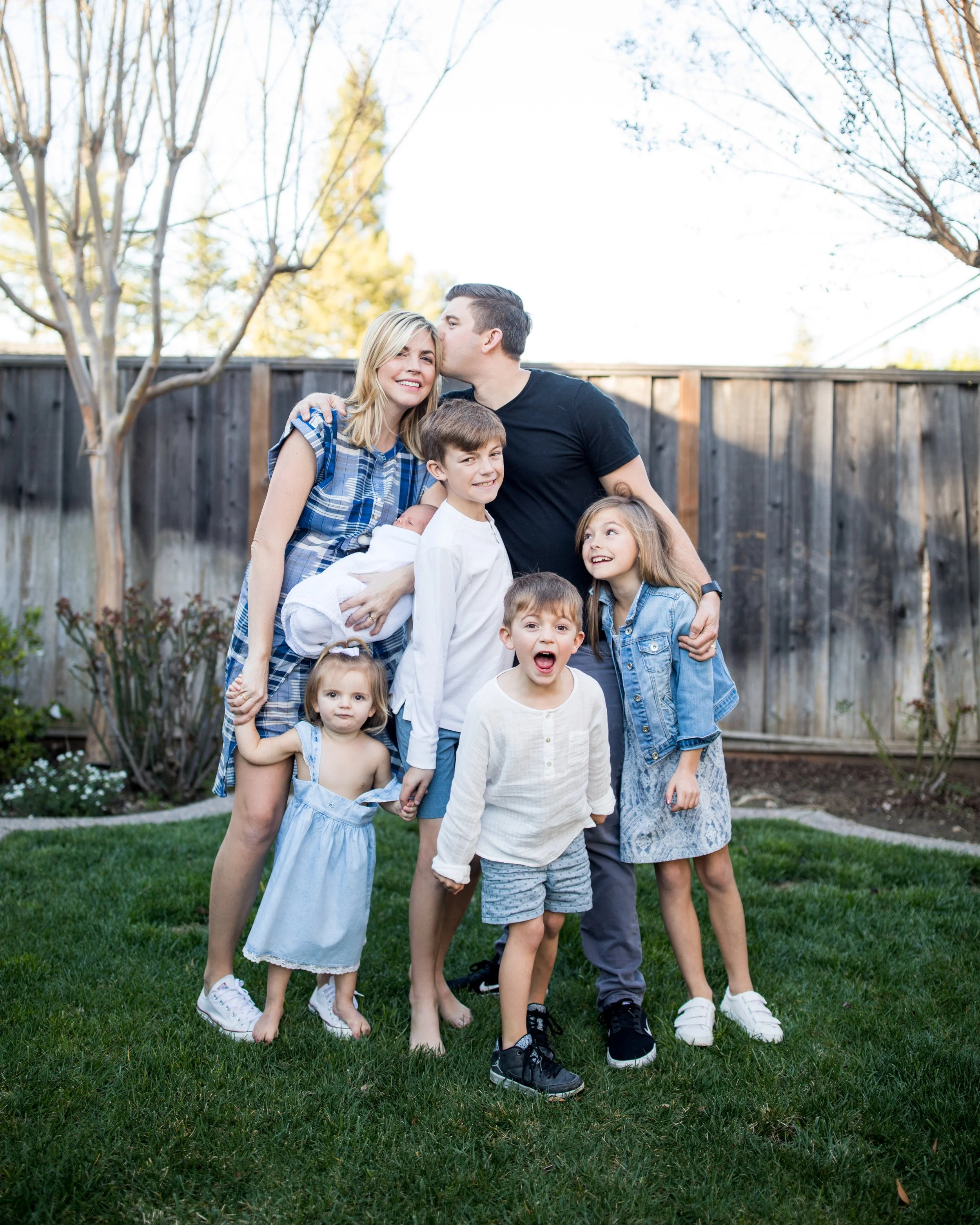 Family of nine with one newborn outside in a backyard, group hugging and smiling near a wooden fence, trees, and grass on a sunny day.