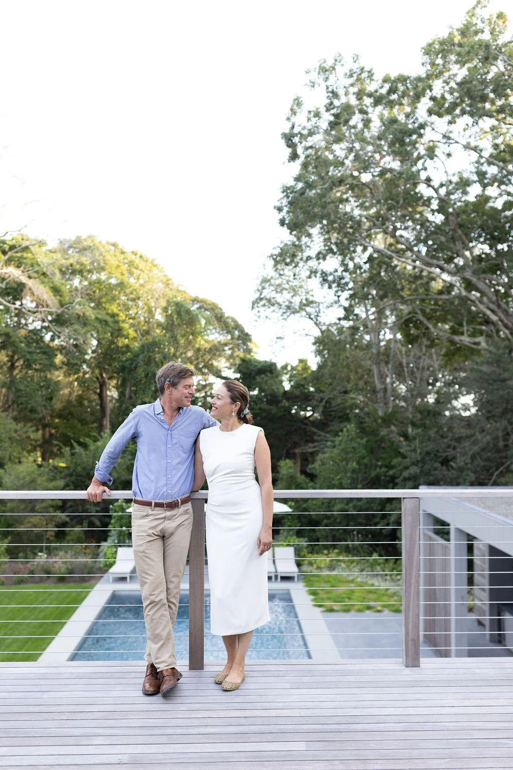 A man and a woman standing on a wooden deck near a swimming pool, smiling and looking at each other, with lush green trees in the background.