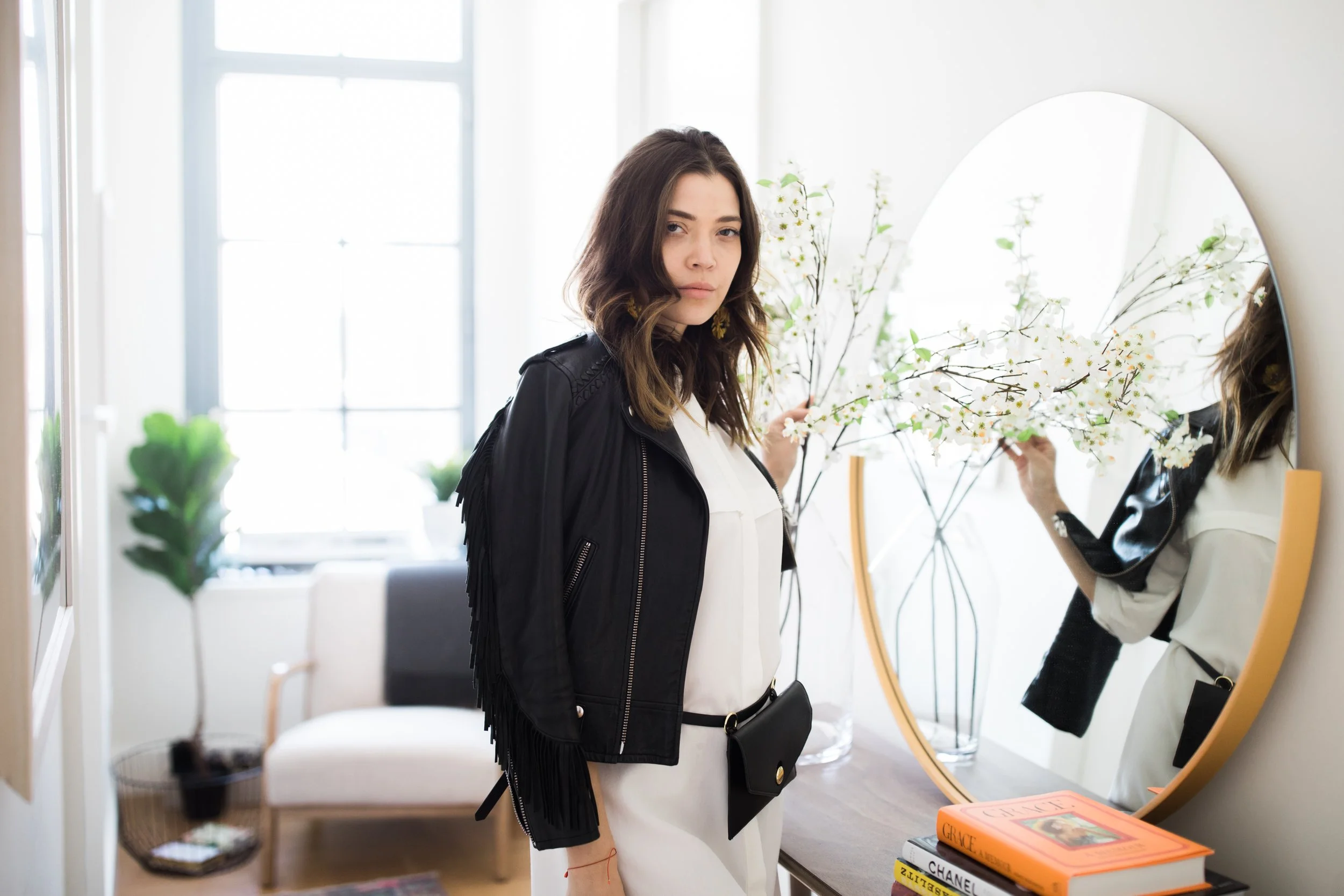 A woman with brown wavy hair looking at herself in a mirror, holding a branch with white flowers. She is wearing a black jacket and a white top inside a bright room with large windows, a potted plant, and a stack of books on a table.