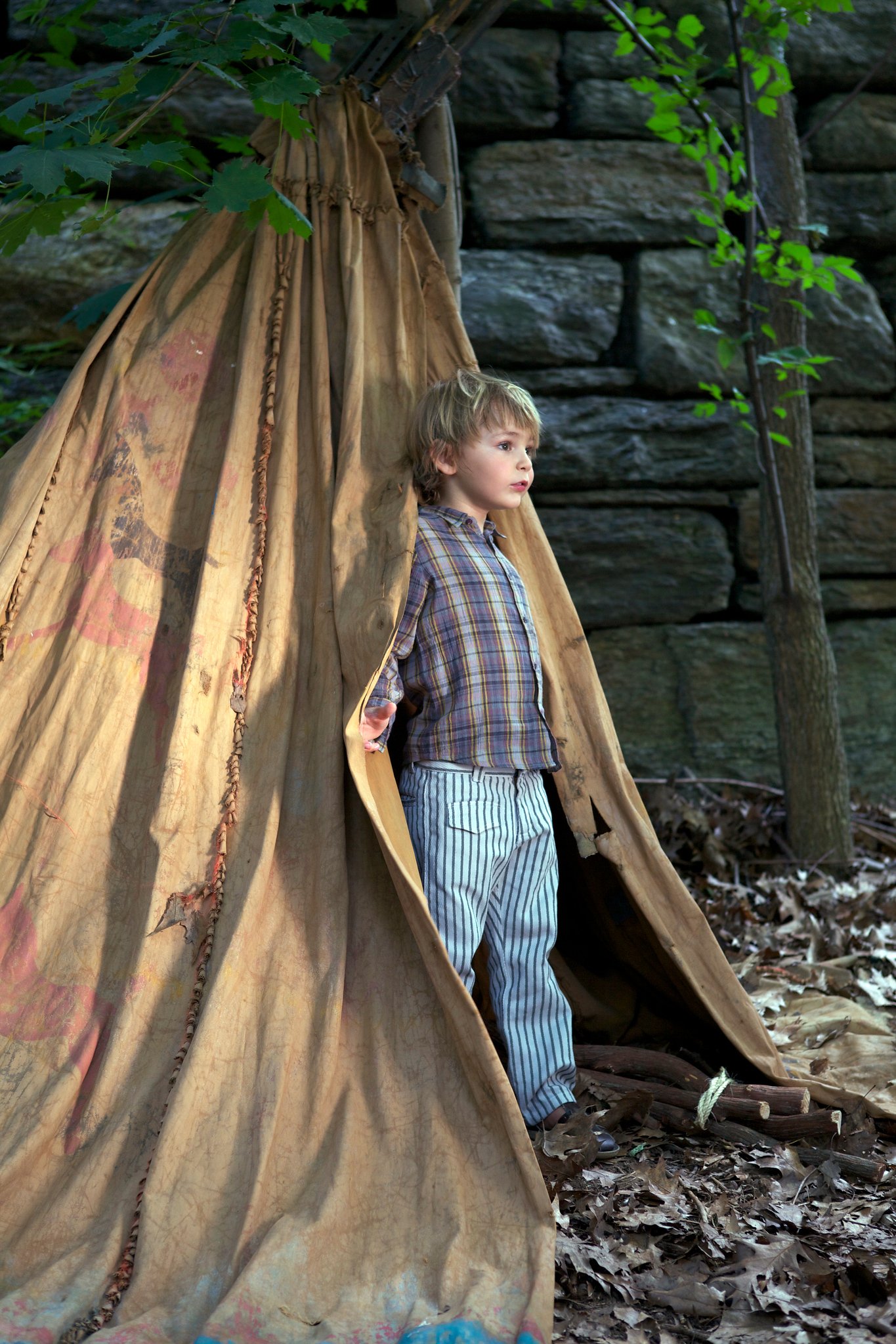 A young boy with blonde hair and striped pants standing inside a beige teepee made of fabric in a wooded area with green leaves and rocks in the background.