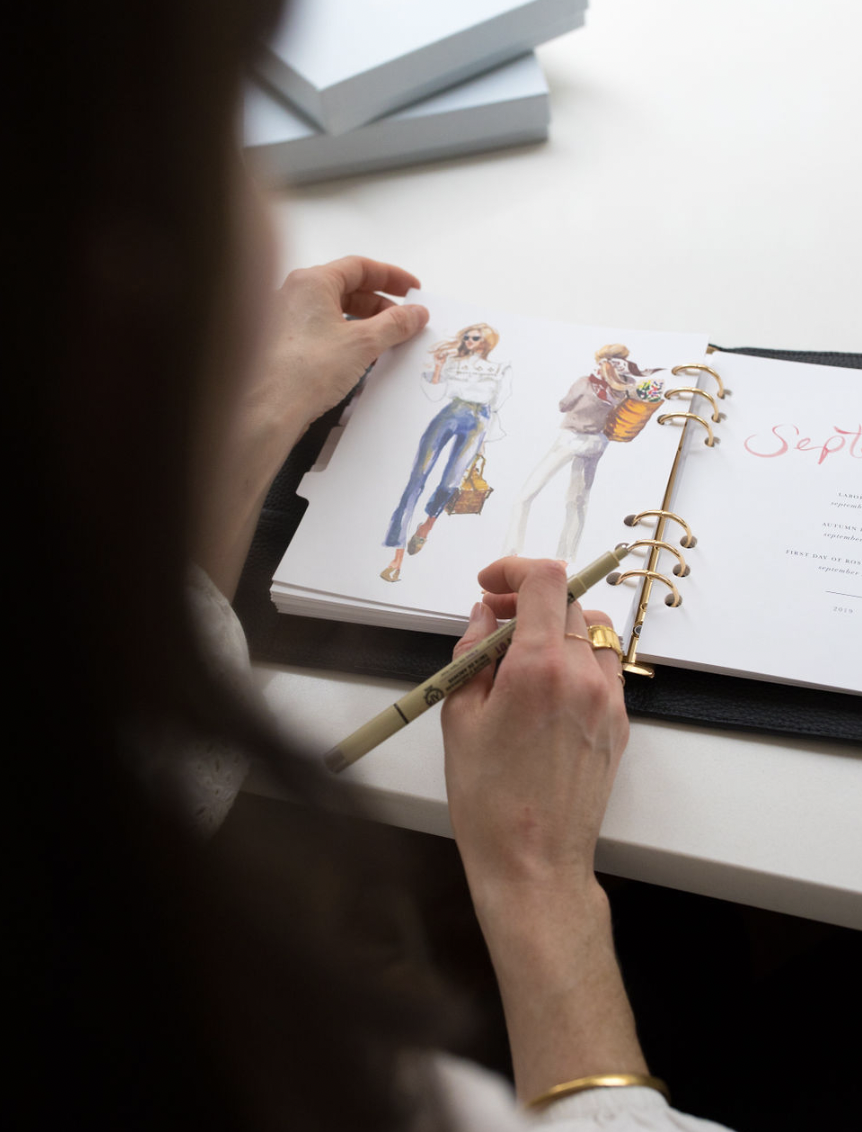 A person with long dark hair, wearing a white shirt, is sitting at a white desk and working in a planner. The planner is open to a page with the month of September, featuring fashion illustrations of two women. The person is holding a gold pen and ha
