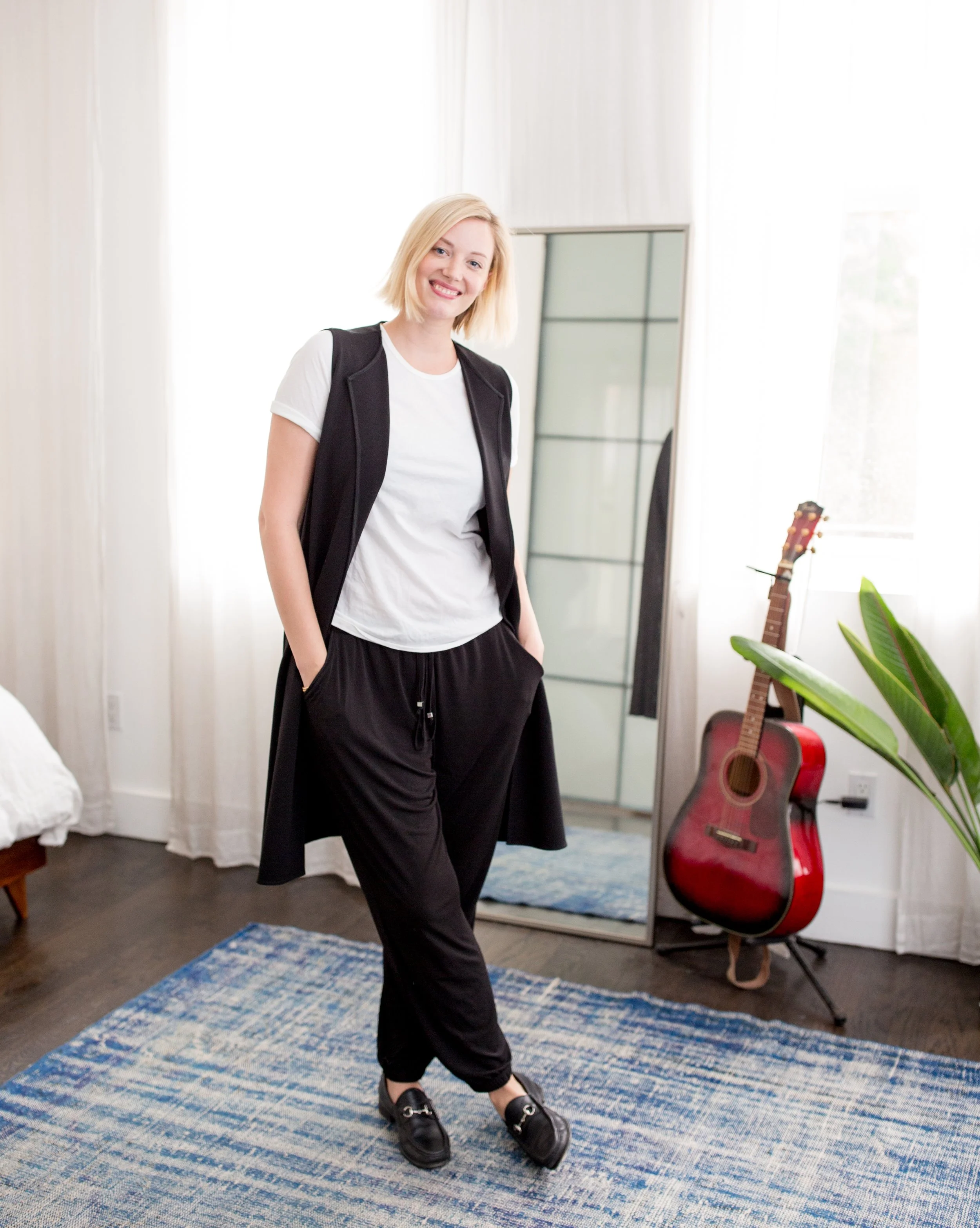 A woman with Blonde hair and a white T-Shirt, a black vest, and black pants standing in a room with natural light, a blue patterned rug, a guitar, a mirror, and houseplants.