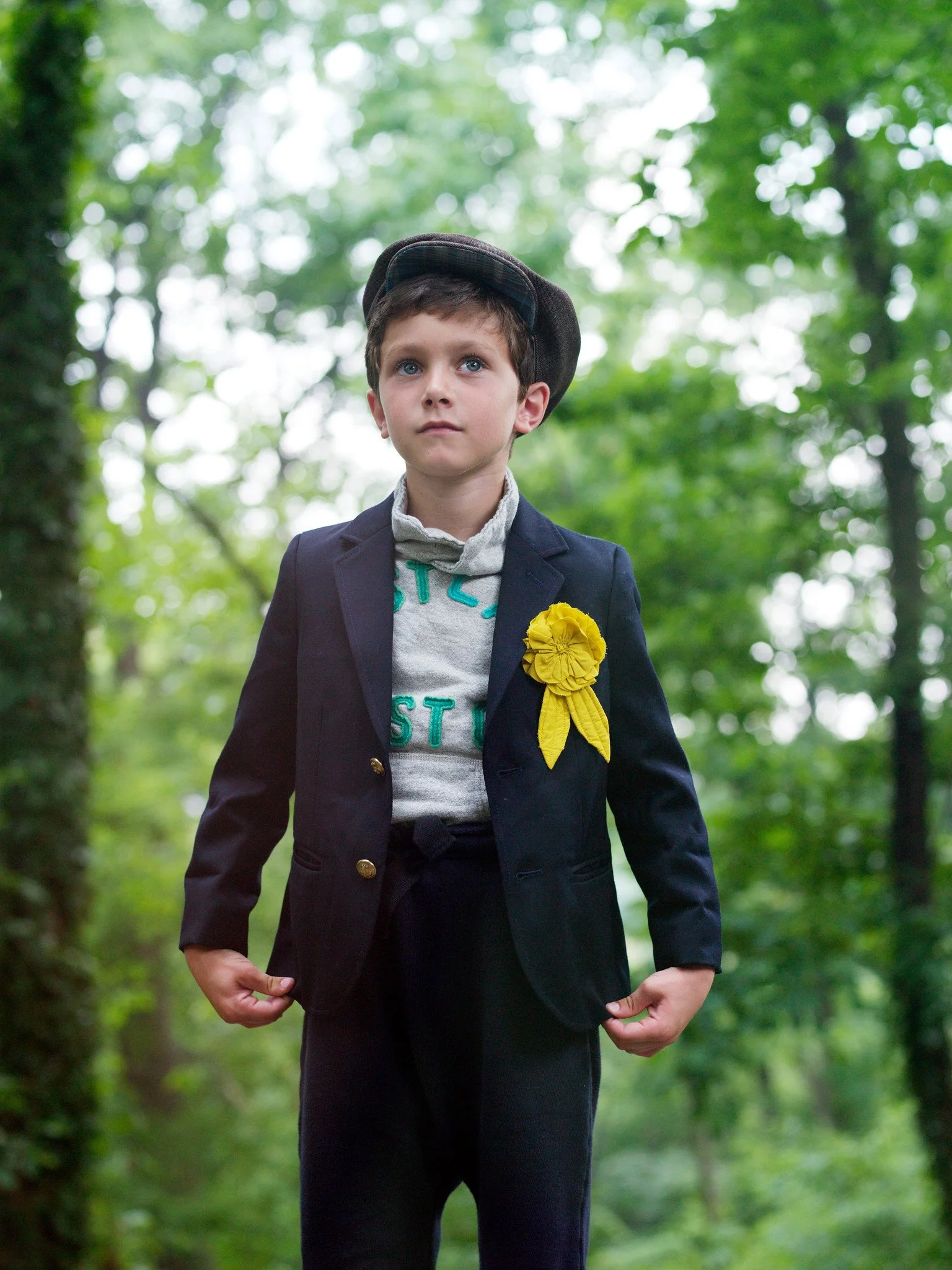 Young boy dressed in vintage-style clothing, including a blazer, holding a yellow flower, and standing in a lush green forest.
