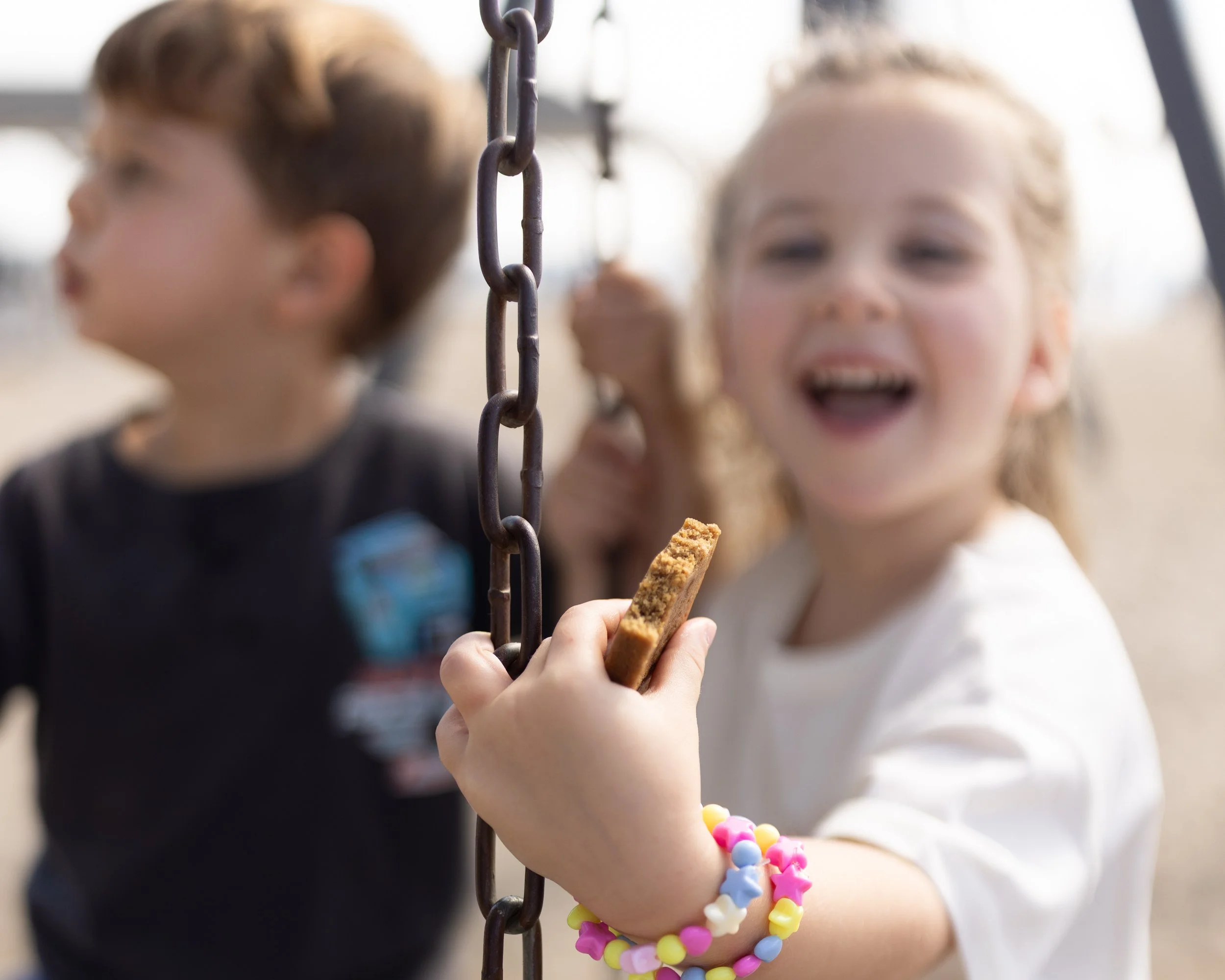Two children holding a cookie and a beaded bracelet, with a metal chain and blurred background.