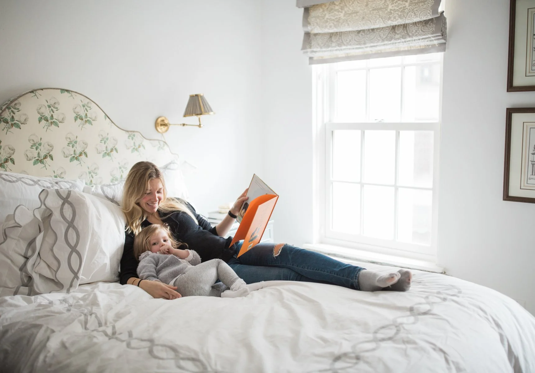 A woman and a young girl relaxing on a bed in a bright, white room, reading a book together.