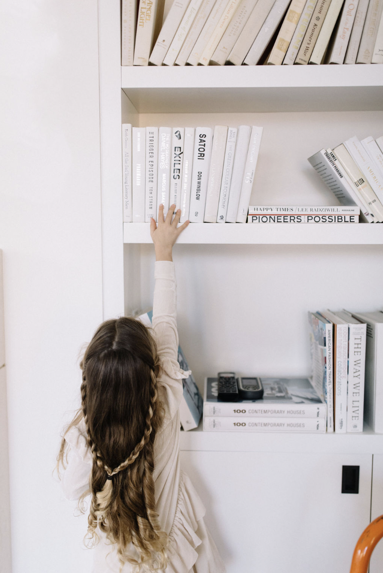 A young girl with long, braided hair reaching up to grab a book from a white bookshelf filled with mostly white and neutral-colored books.