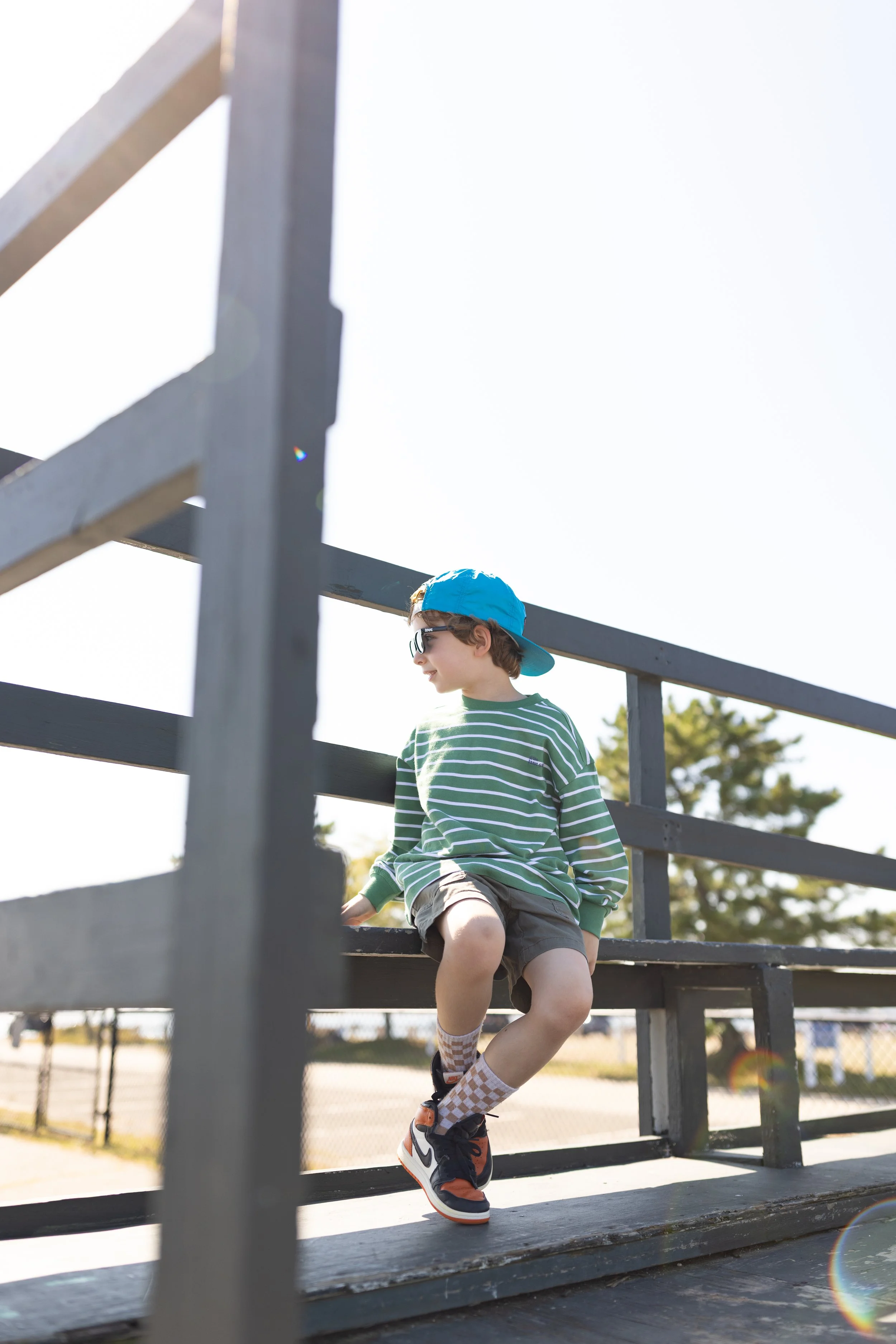 A young boy with a blue backwards baseball cap, sunglasses, a green and white striped shirt, and shorts sitting on a park bench under a clear sky with trees in the background.