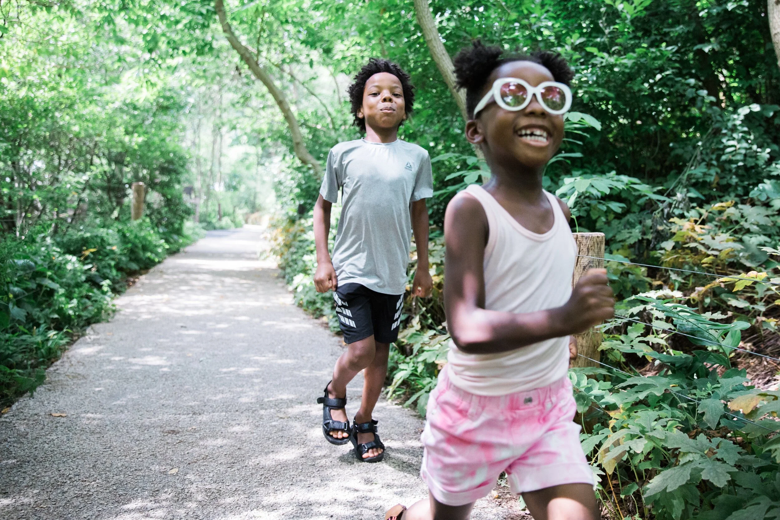 Two children are running along a forest trail with lush green trees and bushes on both sides. The girl in the foreground is smiling and wearing sunglasses, while the boy in the background is looking ahead with a neutral expression.