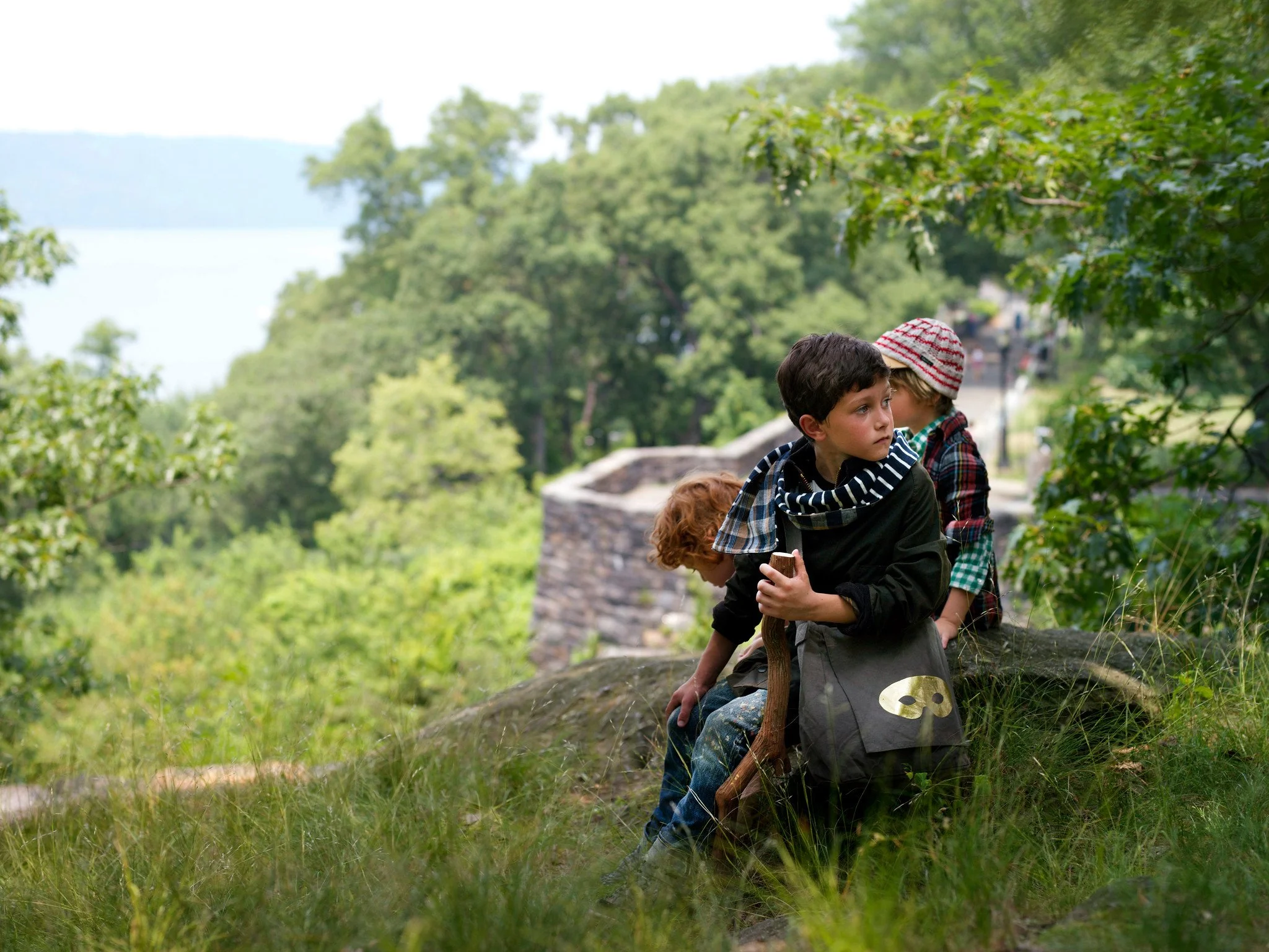 Two young boys sitting outdoors on a rocky ledge near trees and water, one wearing a striped hoodie and the other wearing a red and white beanie.