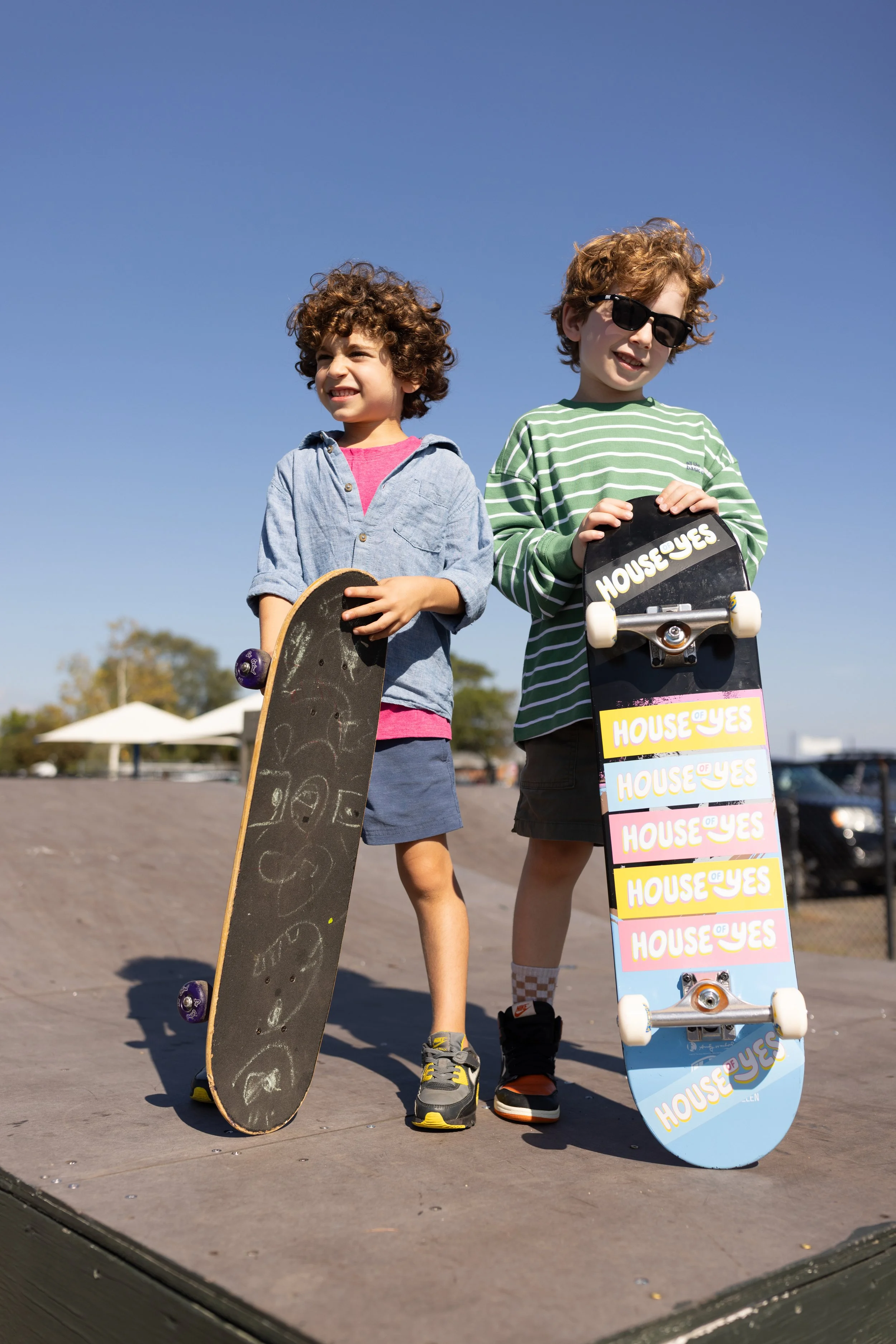 Two young boys standing on a skate ramp holding skateboards outdoors under a clear blue sky. One boy wears sunglasses and a green striped shirt, and the other wears a denim shirt. There are white tents in the background.