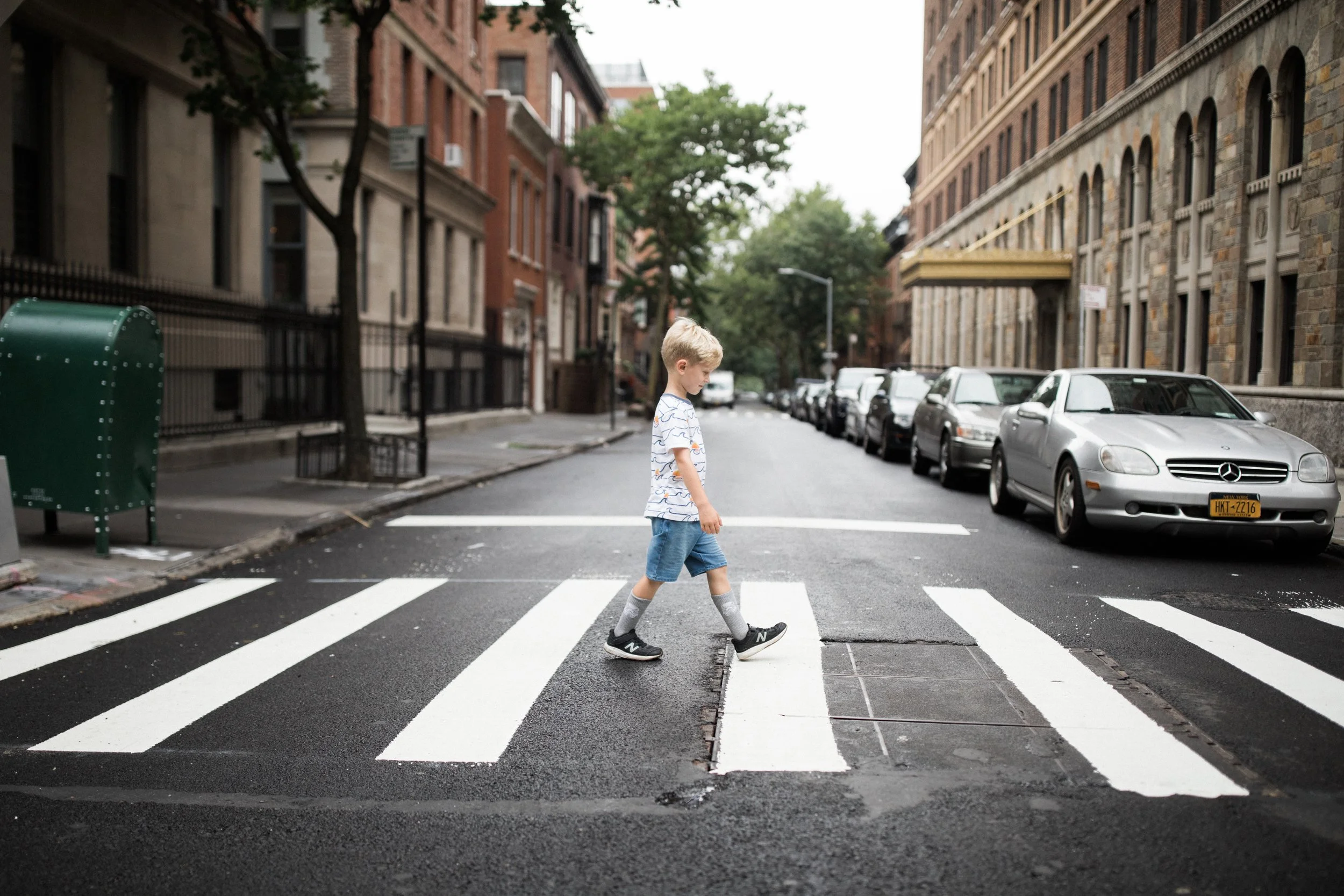 A young boy with blond hair wearing a patterned t-shirt and shorts crossing a street at a crosswalk in an urban neighborhood with parked cars and multi-story brick buildings.
