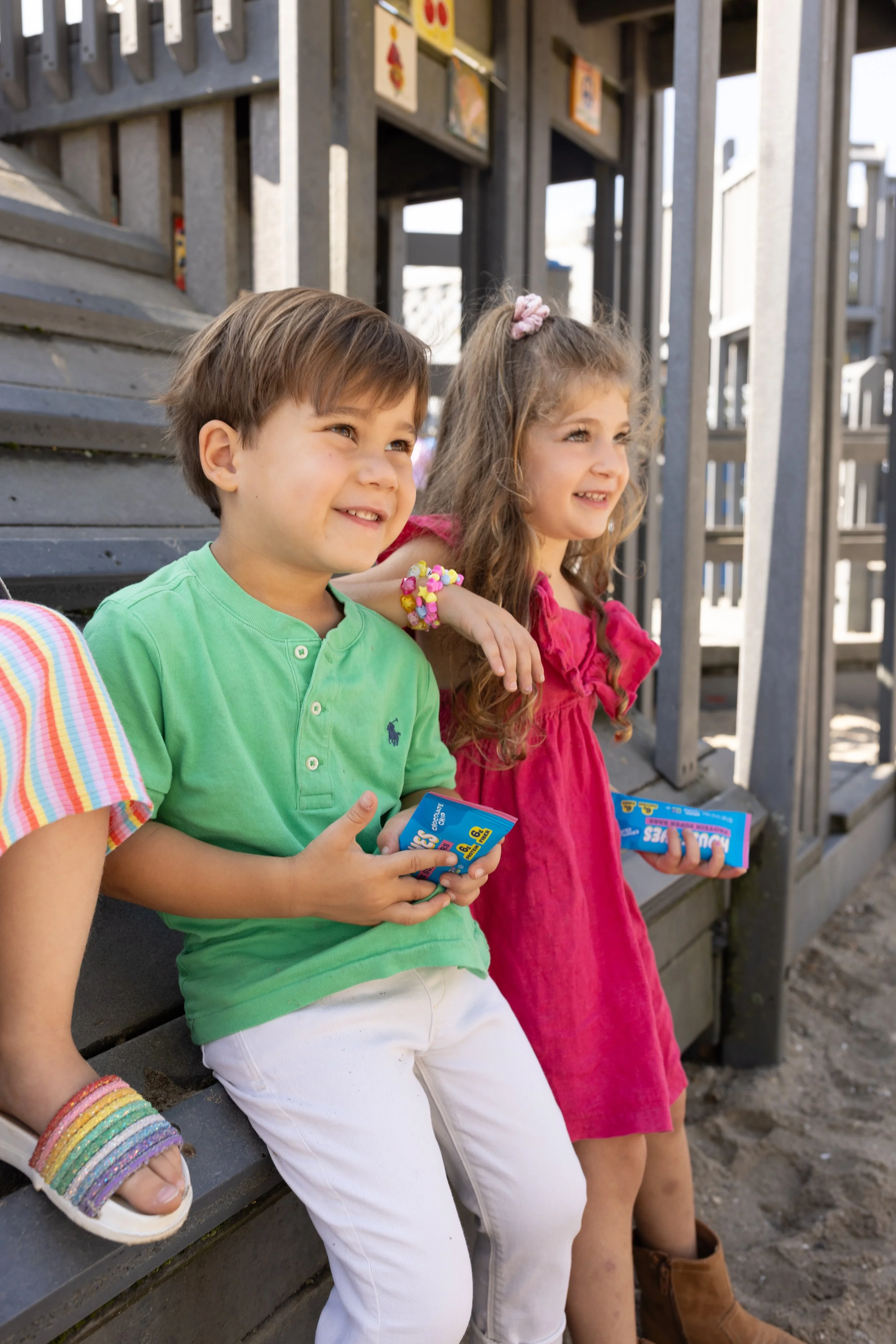 Two young children sitting on a park bench, smiling and holding snack bars. The boy is wearing a green shirt and white pants, while the girl is wearing a pink dress. They are outdoors near a wooden structure with some signs and decorations.