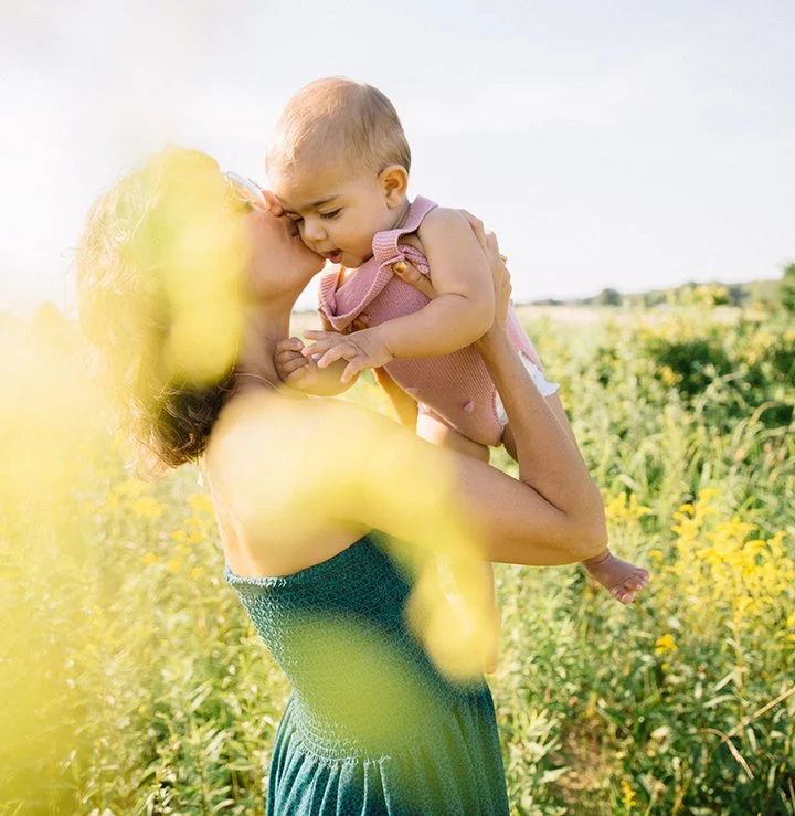 A woman in a green dress holding a baby girl and kissing her in a field of yellow flowers.